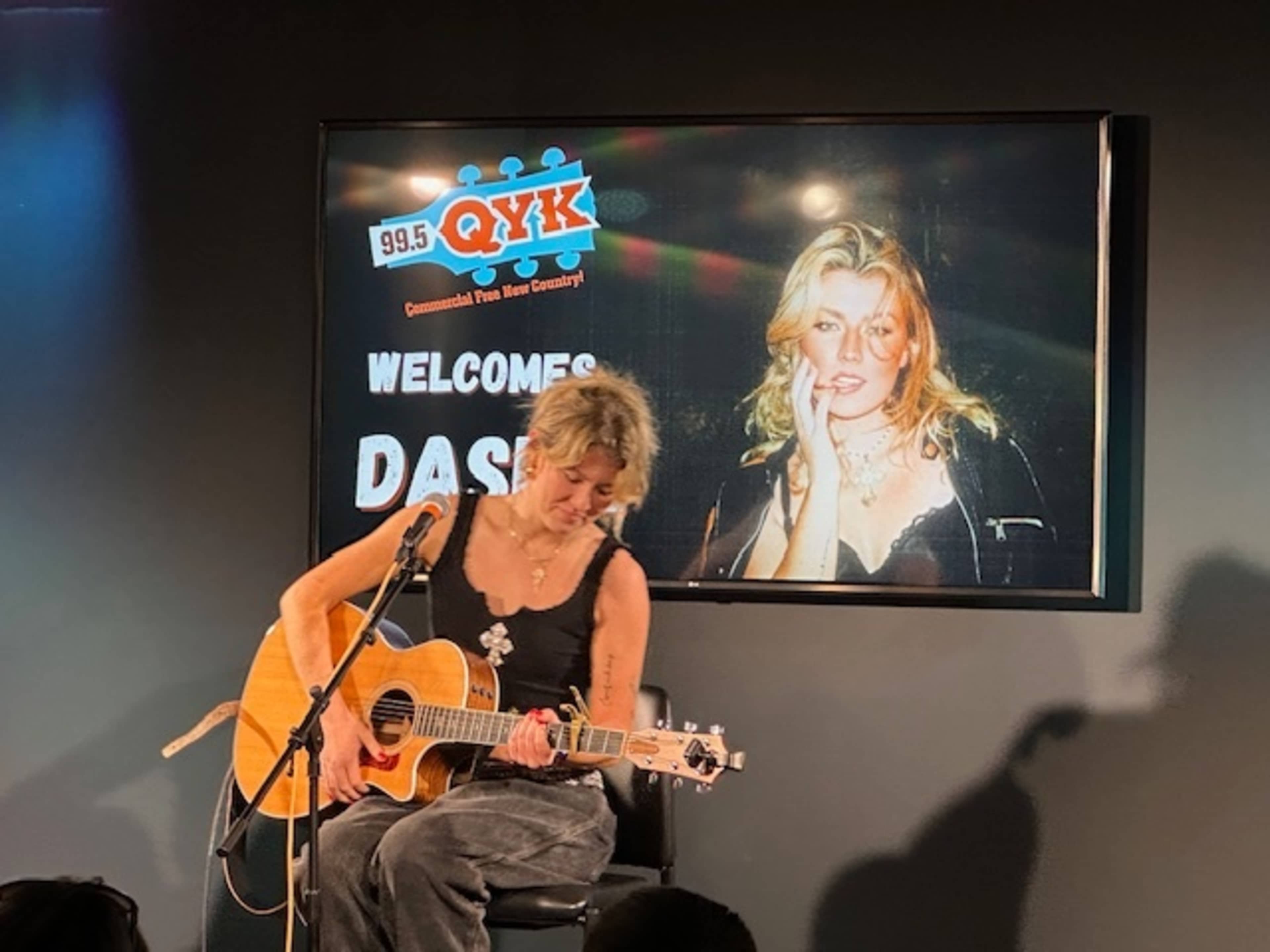 A woman sits on a chair playing an acoustic guitar in front of a screen displaying the text "99.5 QYK Welcomes Dasha."