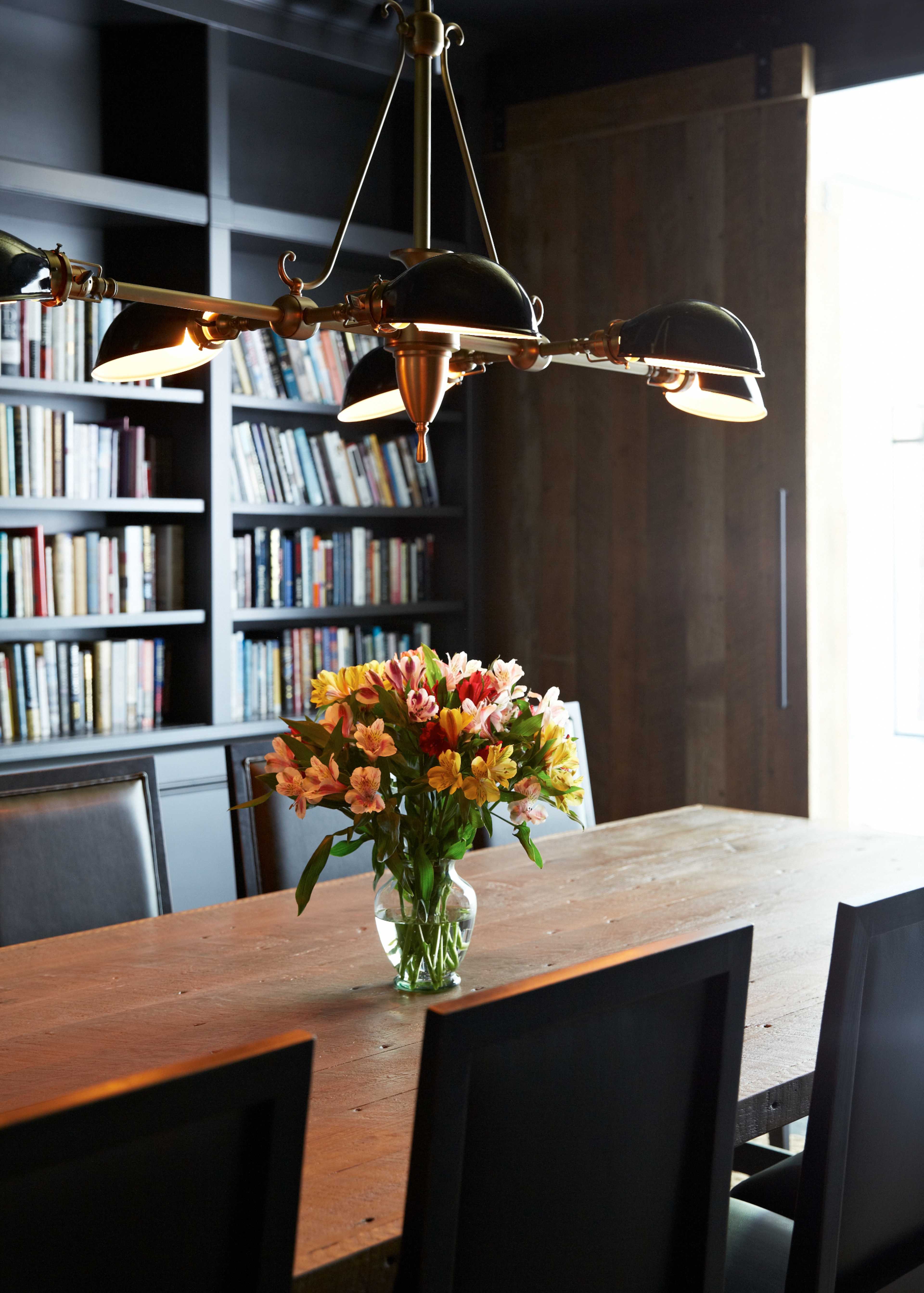 A wooden dining table with a vase of flowers sits under a chandelier in a room lined with bookshelves.