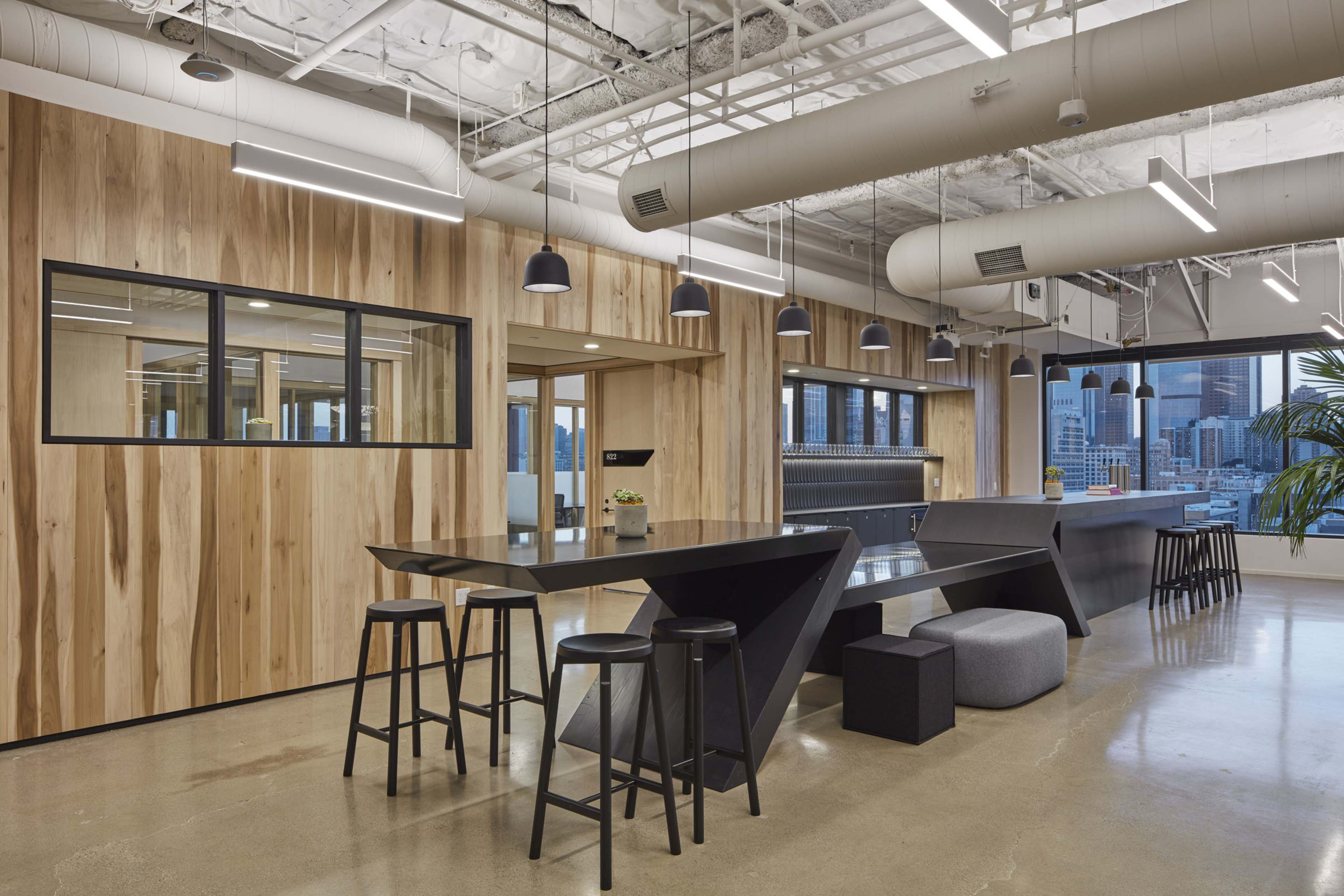The image shows a modern office space featuring wooden paneling, industrial lighting, and a mix of black and gray furniture.