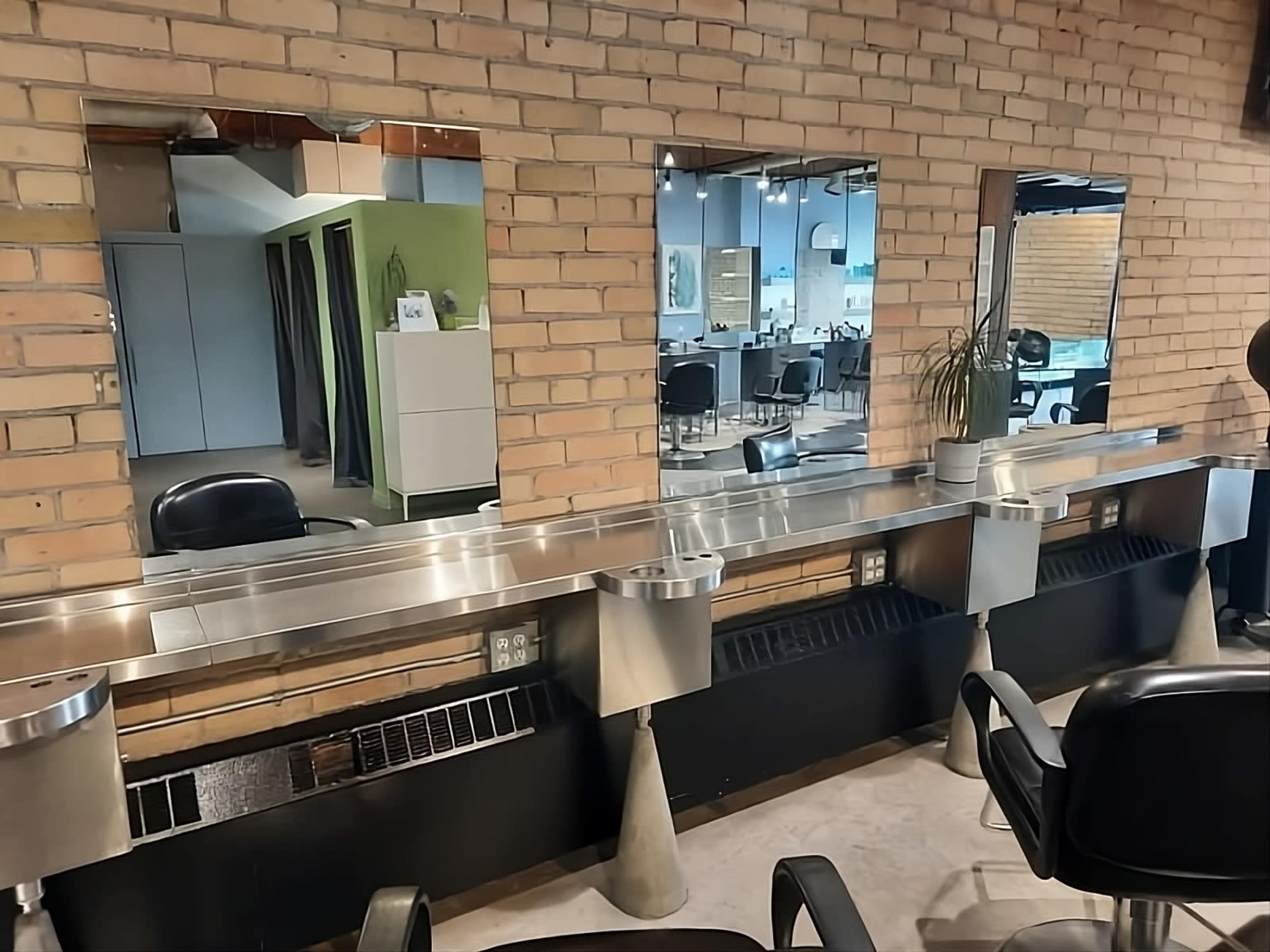 A modern hair salon with three mirrors above a sleek, metal counter and black chairs, set against a brick wall.