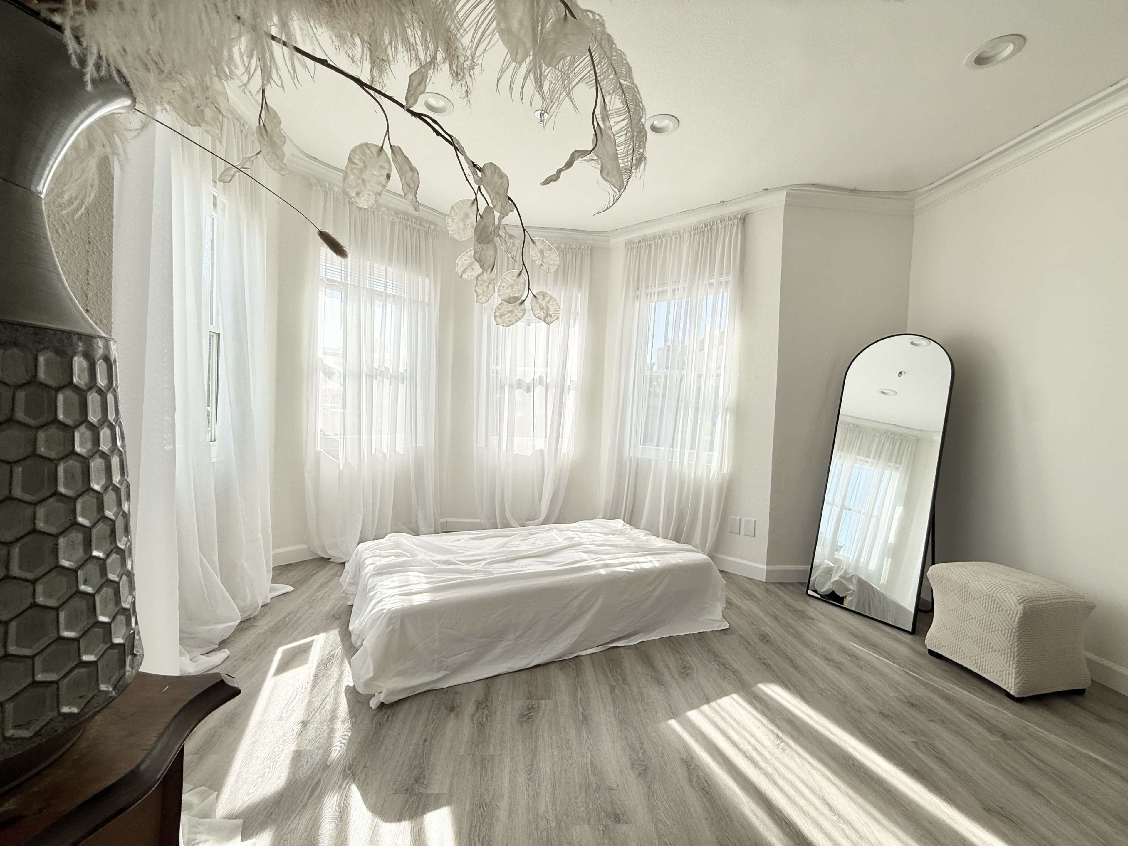 A bright, airy room features a white bed with no bedding, large windows draped in sheer curtains, and a decorative mirror.