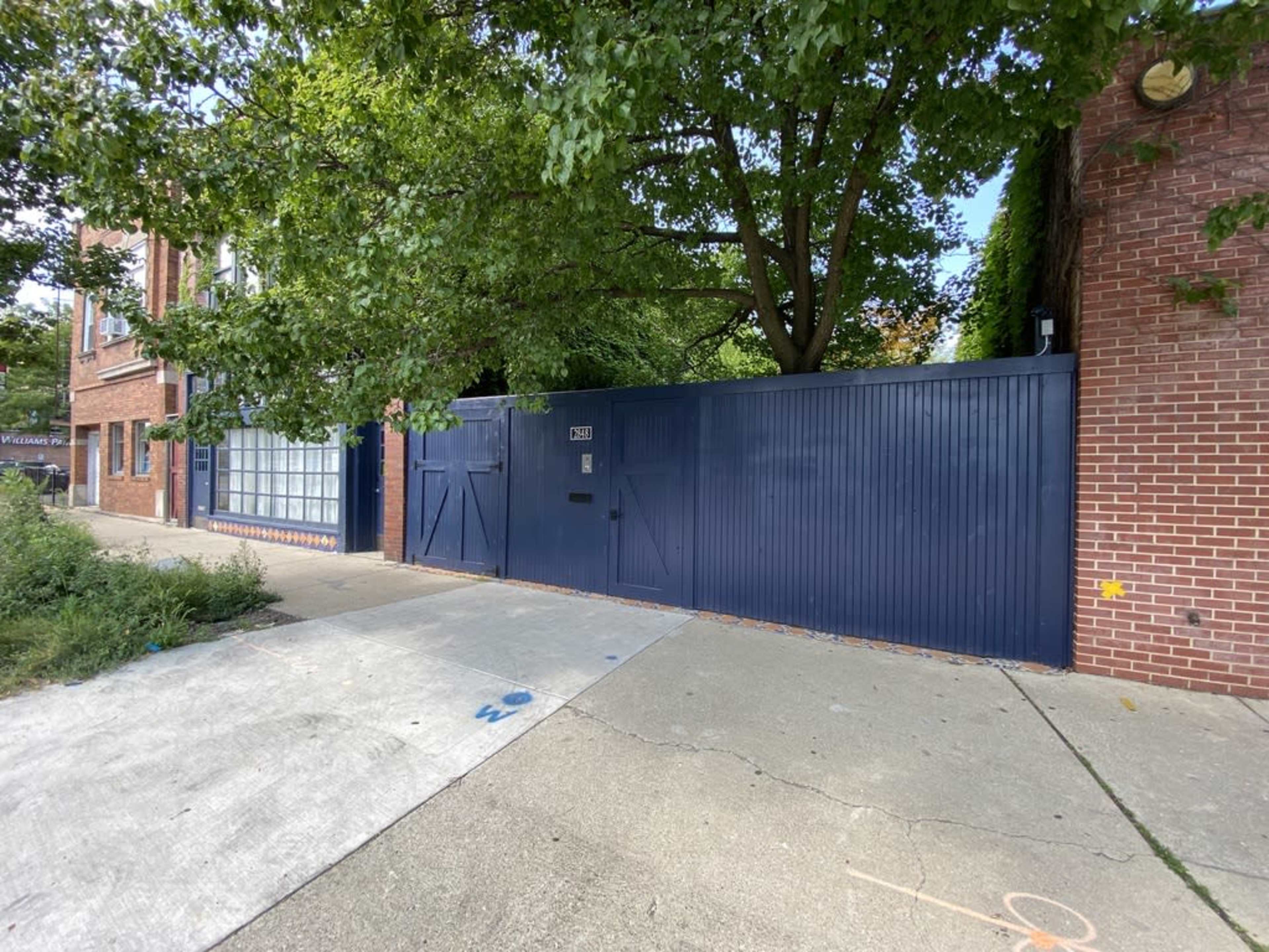 A blue wooden gate is set against a brick building and a large tree in a residential area.