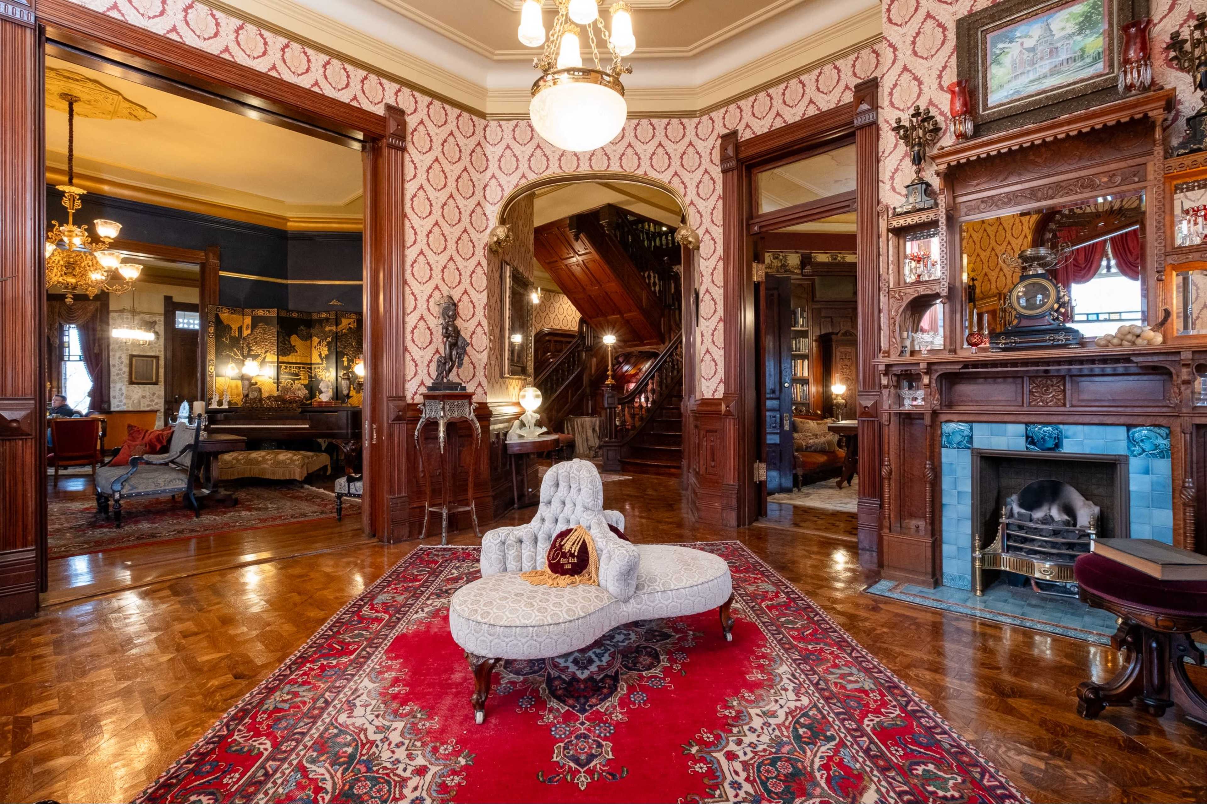 The image shows a richly decorated Victorian-style living room with ornate woodwork, vintage furnishings, and a central chaise lounge on a patterned rug.