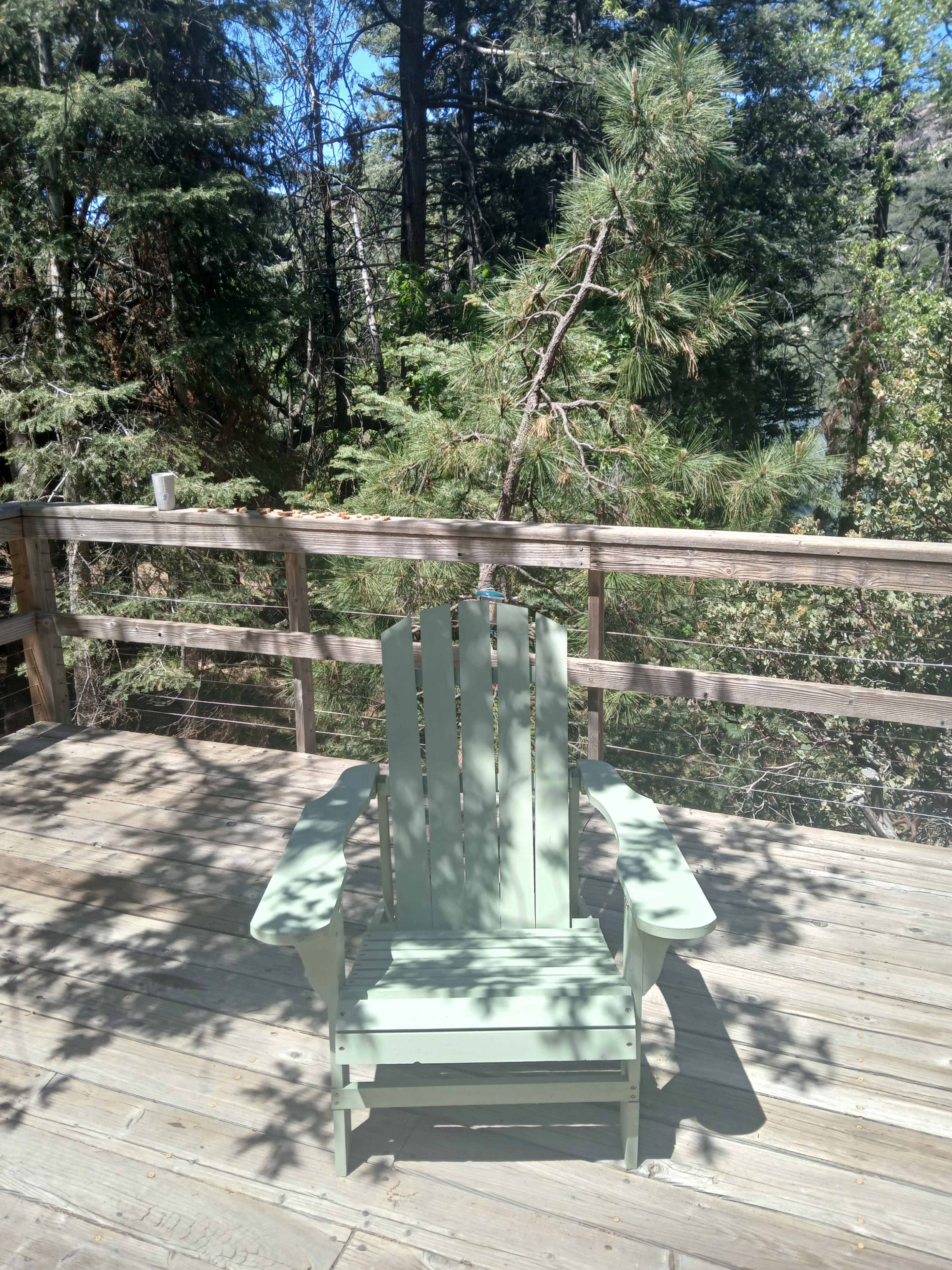 A green Adirondack chair sits on a wooden deck surrounded by tall trees.