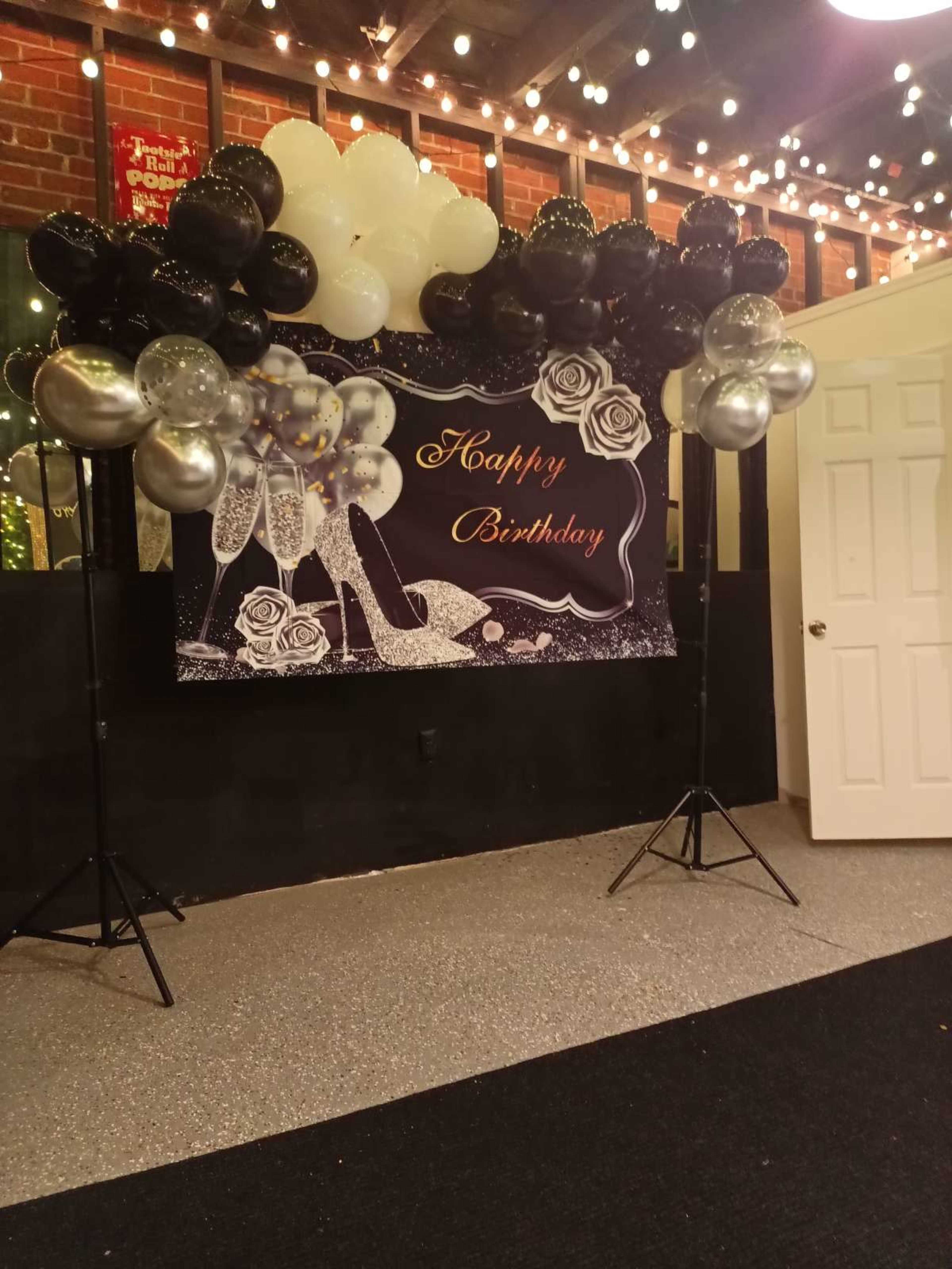 The image features a birthday decoration setup with a "Happy Birthday" banner surrounded by black, white, and silver balloons, under warm string lights.