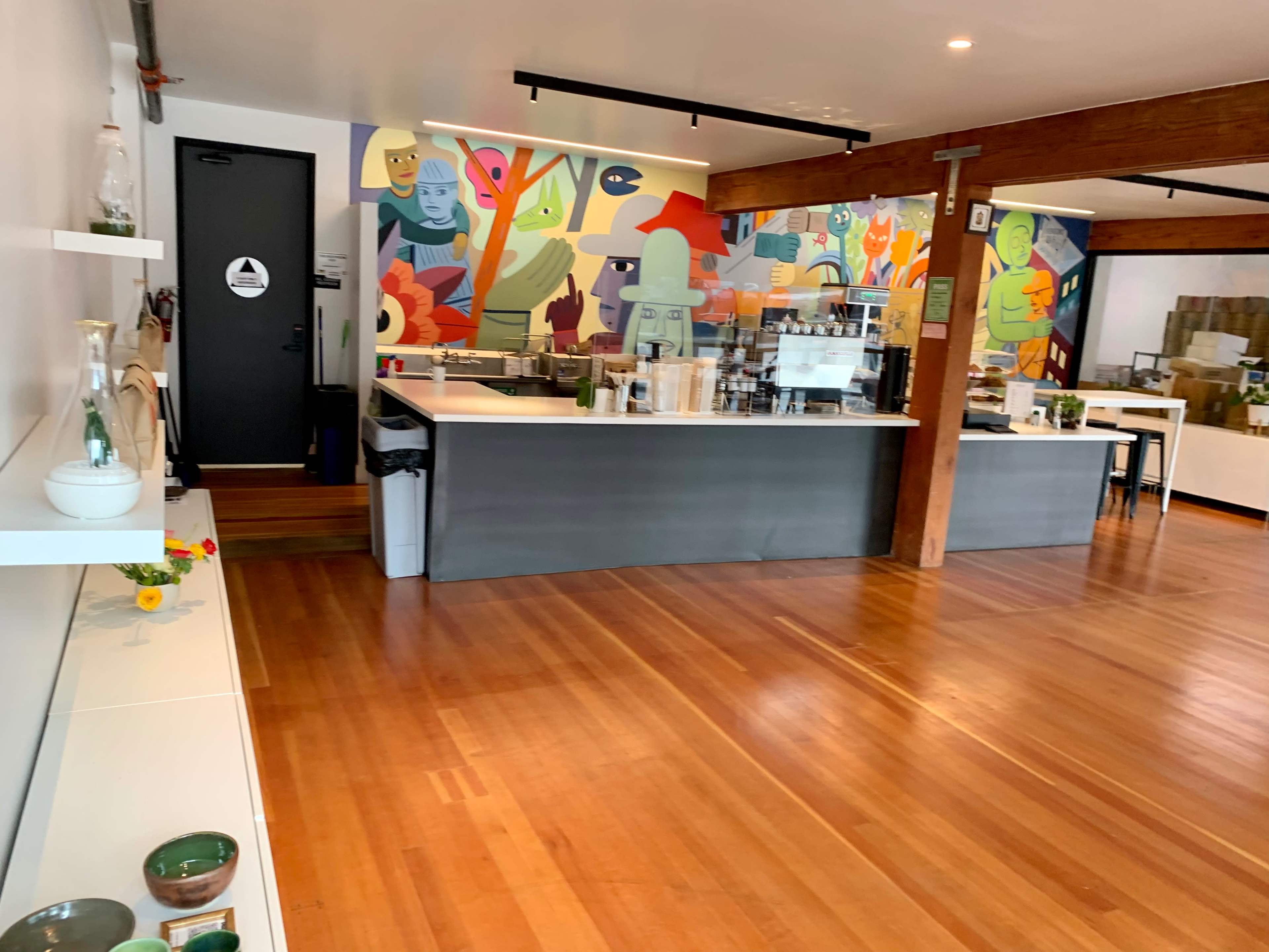 A modern interior of a café featuring a colorful mural, a service counter with a coffee machine, and wooden flooring.