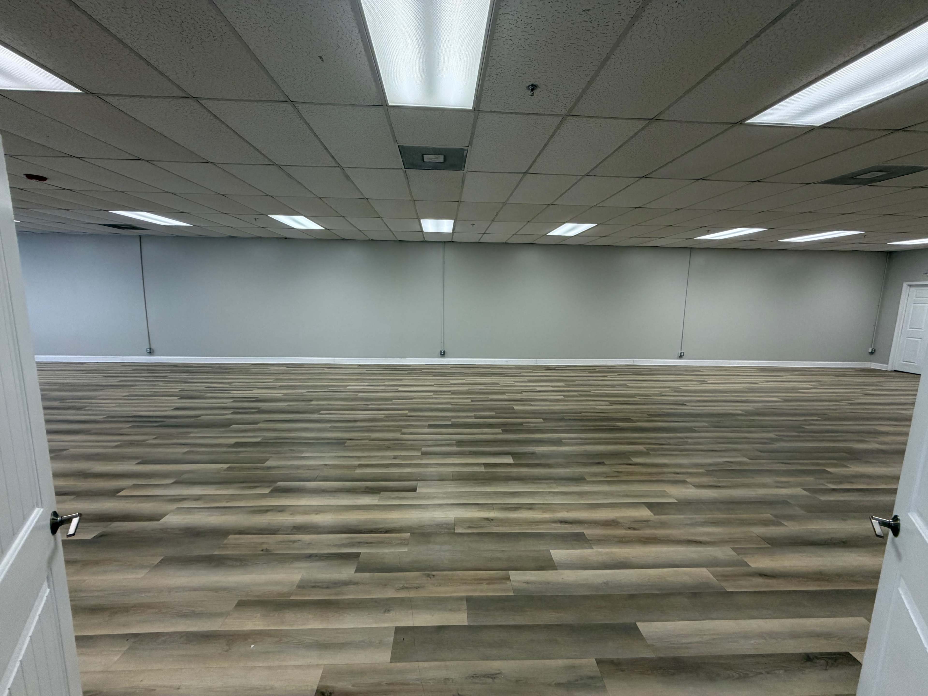 The image shows a spacious, empty room with hardwood flooring and gray walls, illuminated by fluorescent lights.