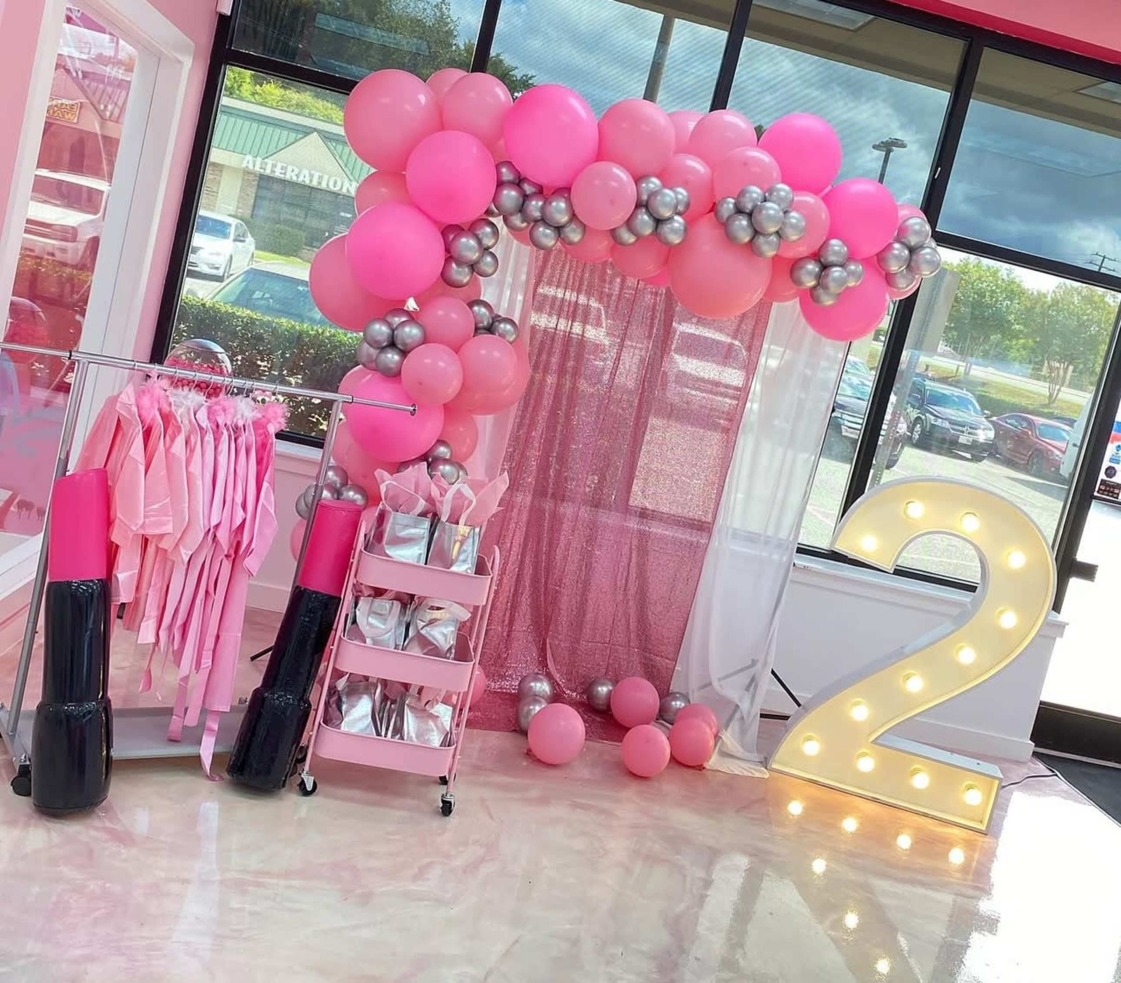 The image shows a festive event area decorated with a balloon arch in pink and silver, a display of clothing, and a large illuminated number 2.