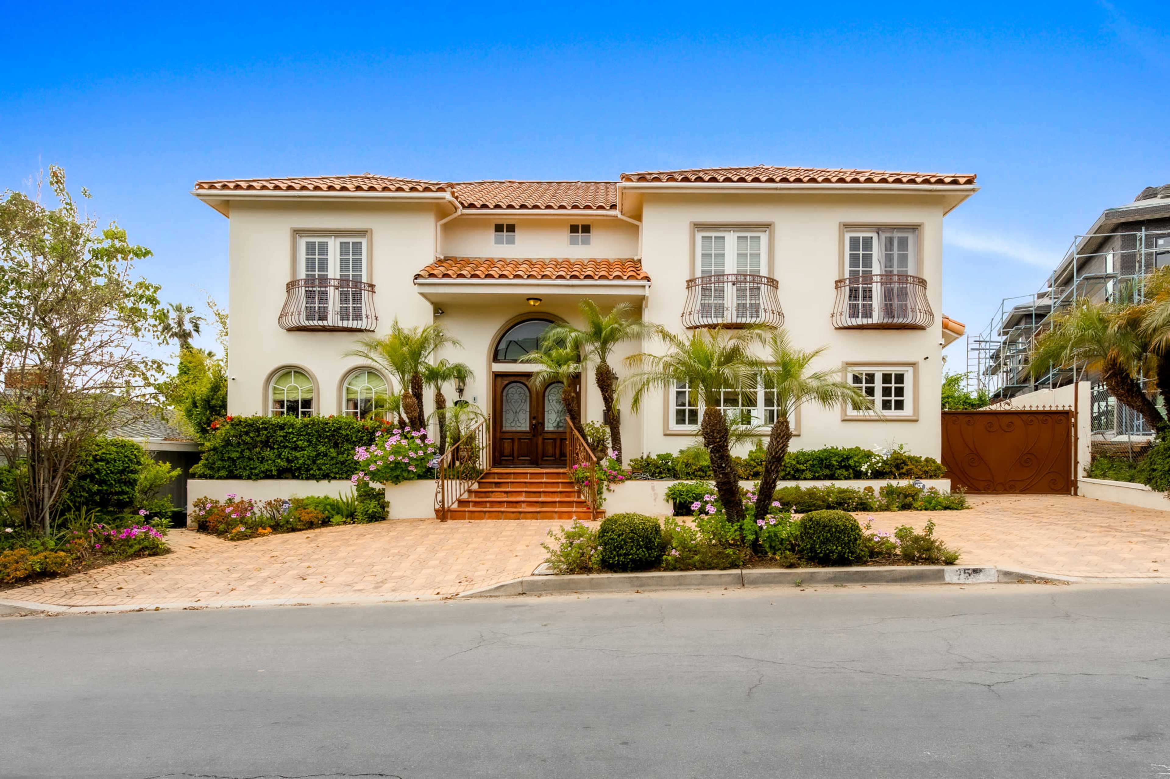 A two-story house with a tiled roof, large front windows, and palm trees is situated on a landscaped lot.
