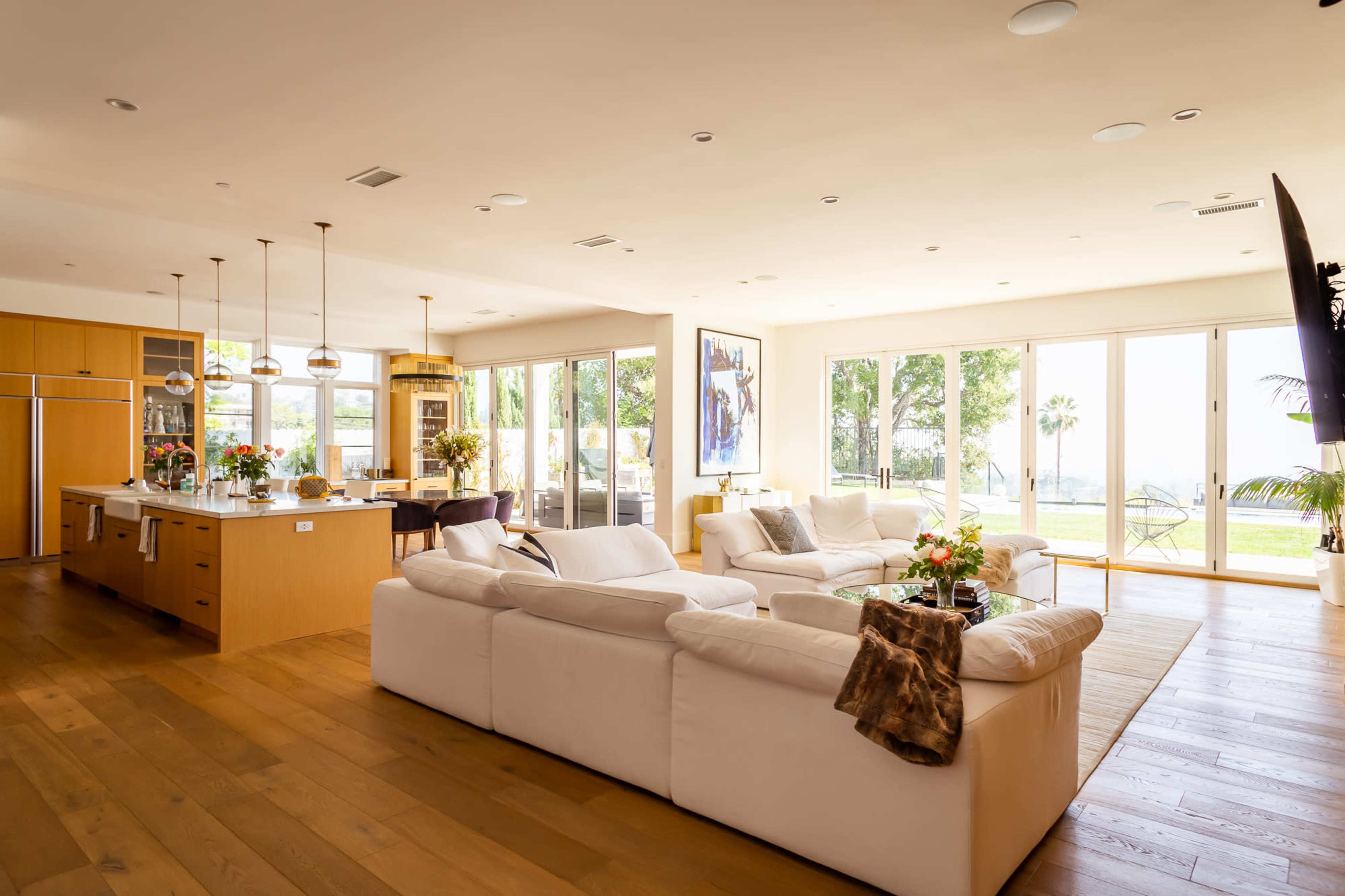 A spacious living room features white sofas and a kitchen area with wooden cabinetry, large windows, and views of an outdoor patio.