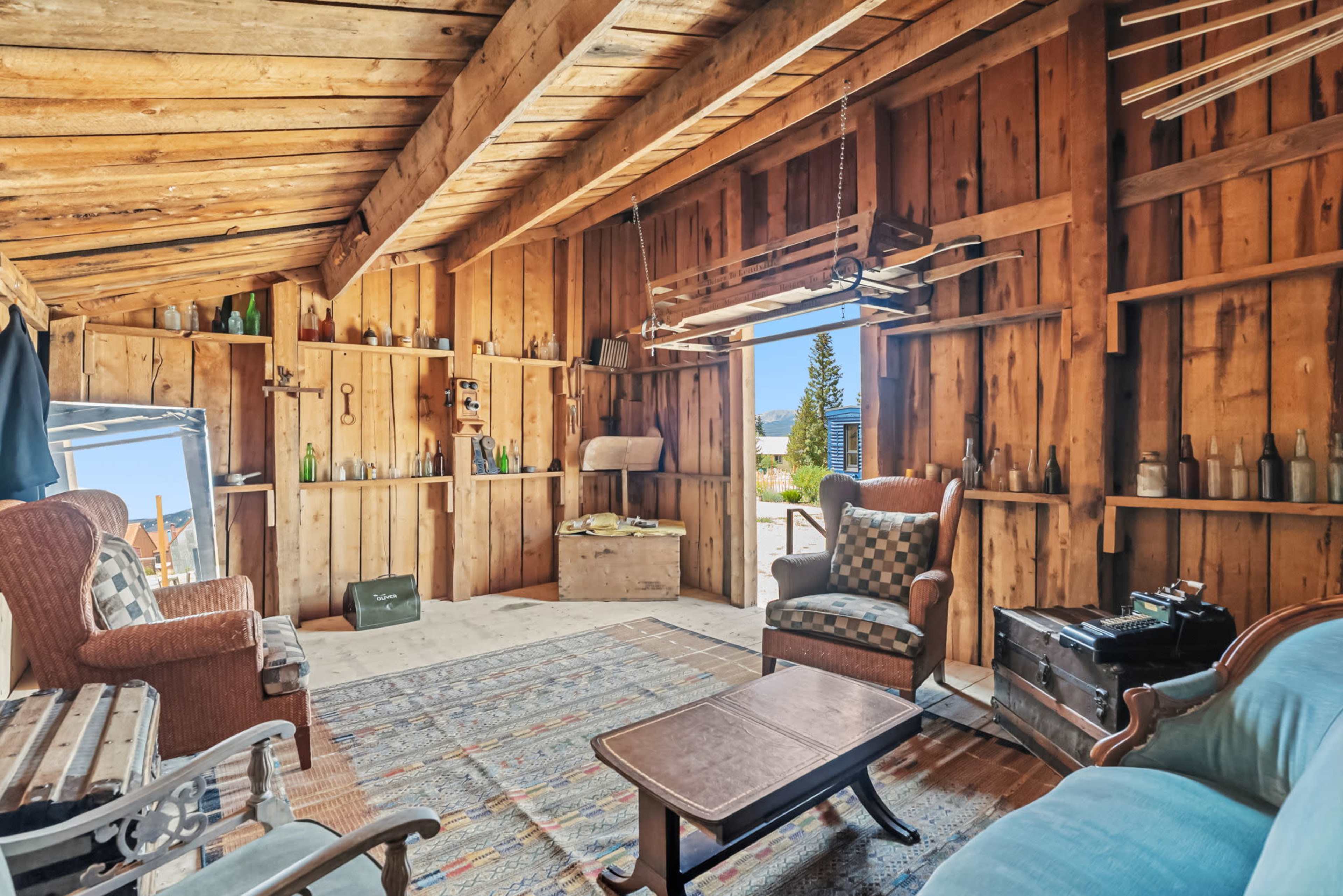 The image shows the interior of a rustic cabin with wooden walls, two chairs, a small coffee table, and a patterned rug.
