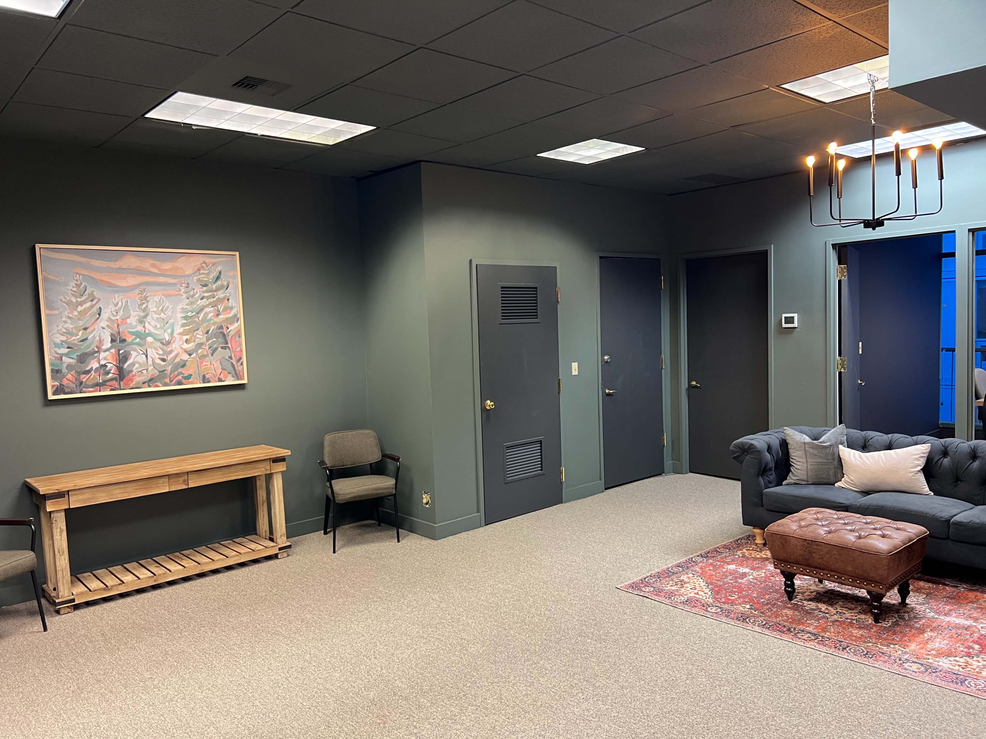 A sparsely furnished office space with a green walls, a framed landscape painting, a wooden console table, a gray sofa, and a brown ottoman on a patterned rug.