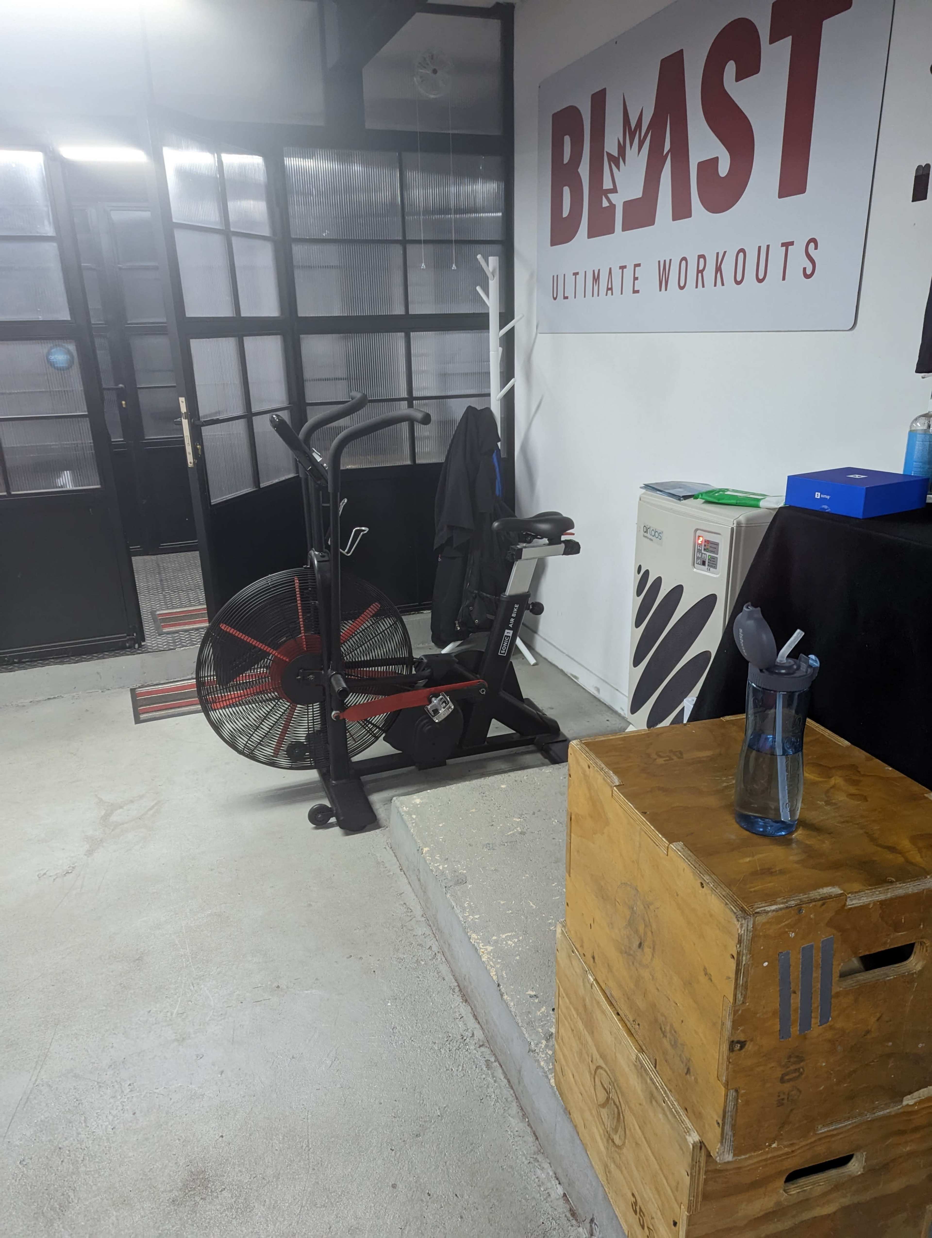 A fitness area with an exercise bike next to wooden boxes and a water bottle on a platform, along with a sign that reads "BLAST ULTIMATE WORKOUTS."