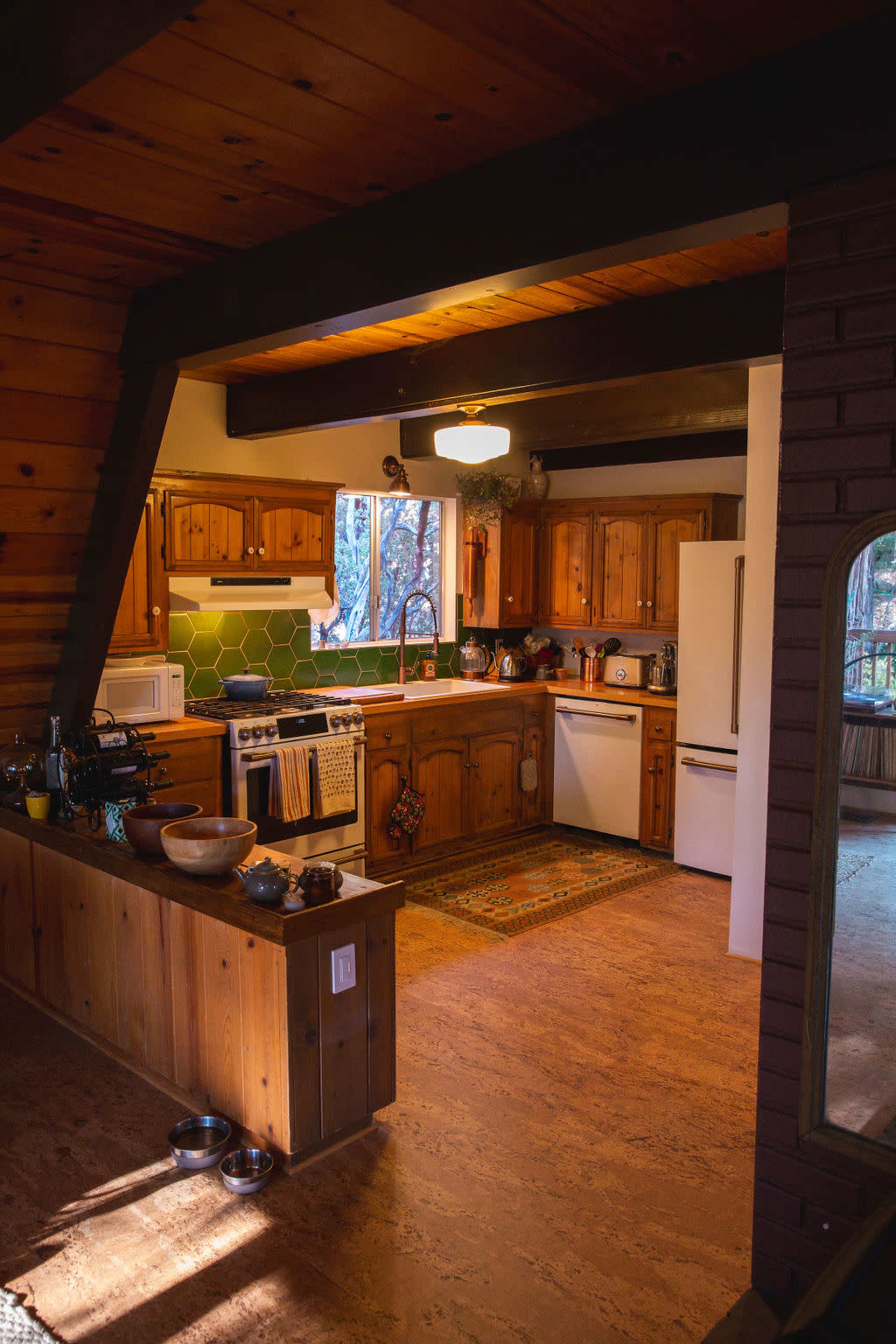 The image shows a wooden kitchen with hexagonal green tiles, natural lighting, and various kitchen appliances and utensils neatly arranged.