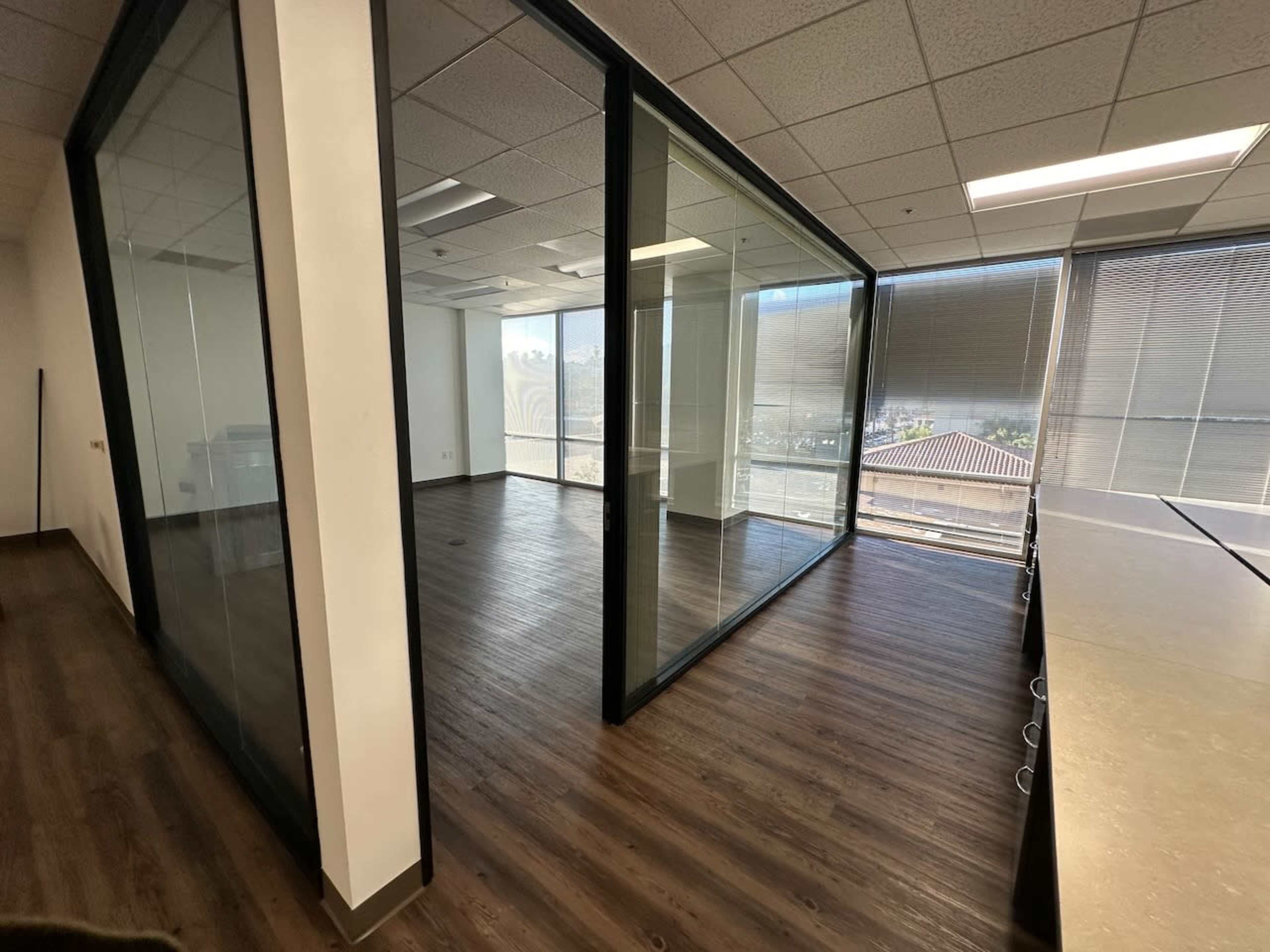 A modern office space featuring a glass-walled meeting room, large windows, and hardwood flooring.