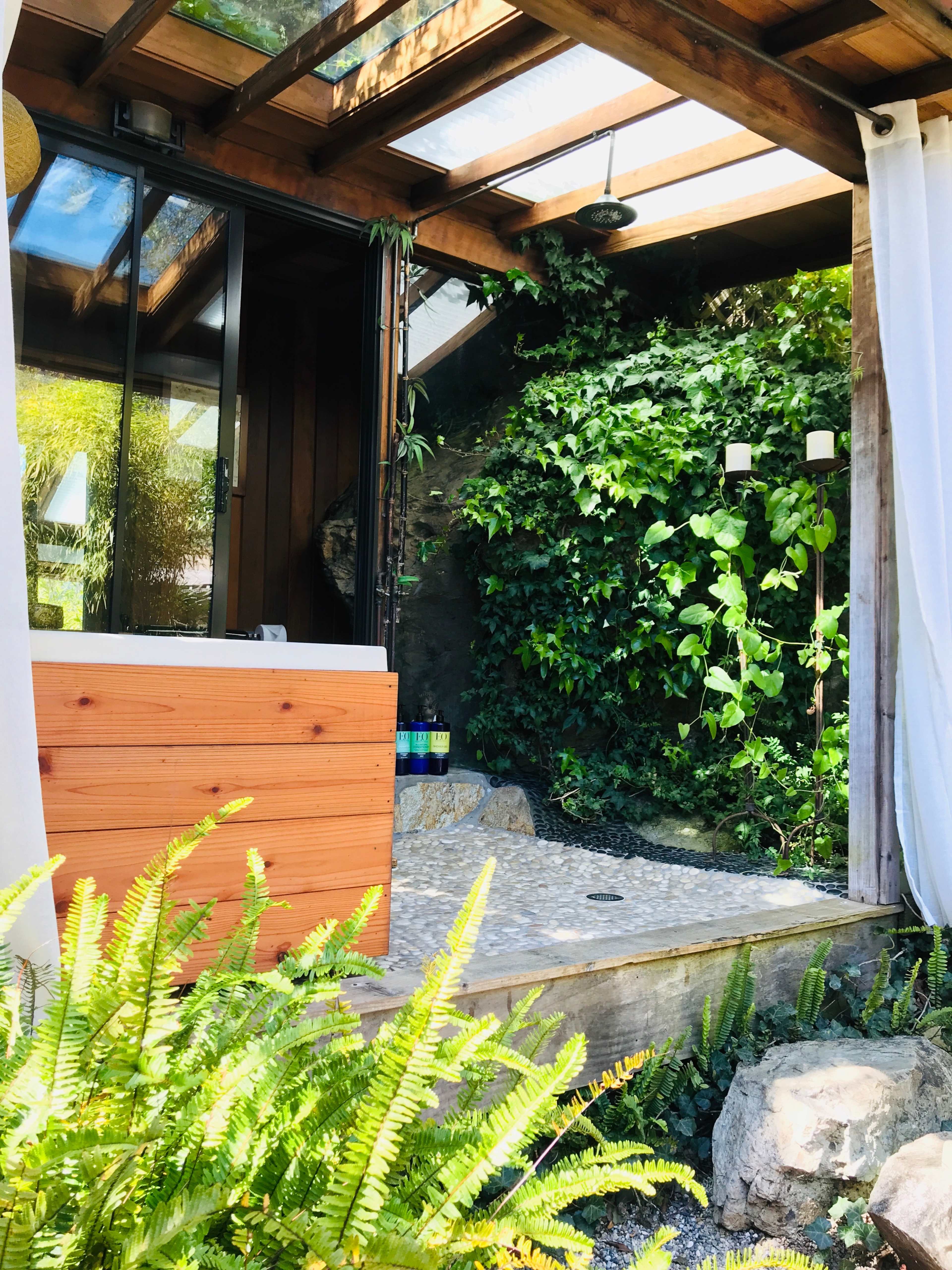 The image shows a wooden structure with large glass windows, a private outdoor shower area, and lush greenery surrounding it.