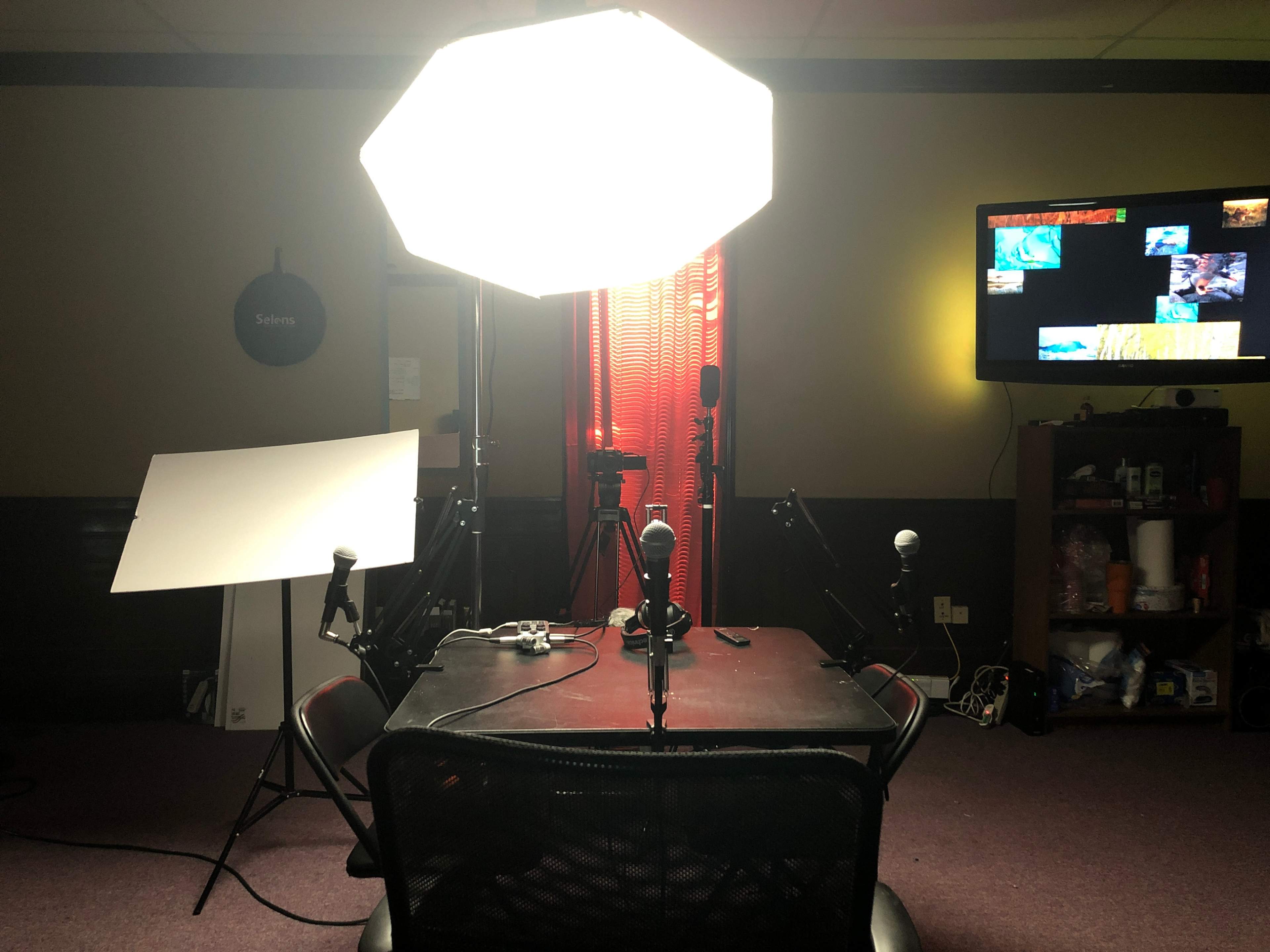 The image shows a recording studio setup with a round table, multiple microphones, bright lights, and a television displaying video content.