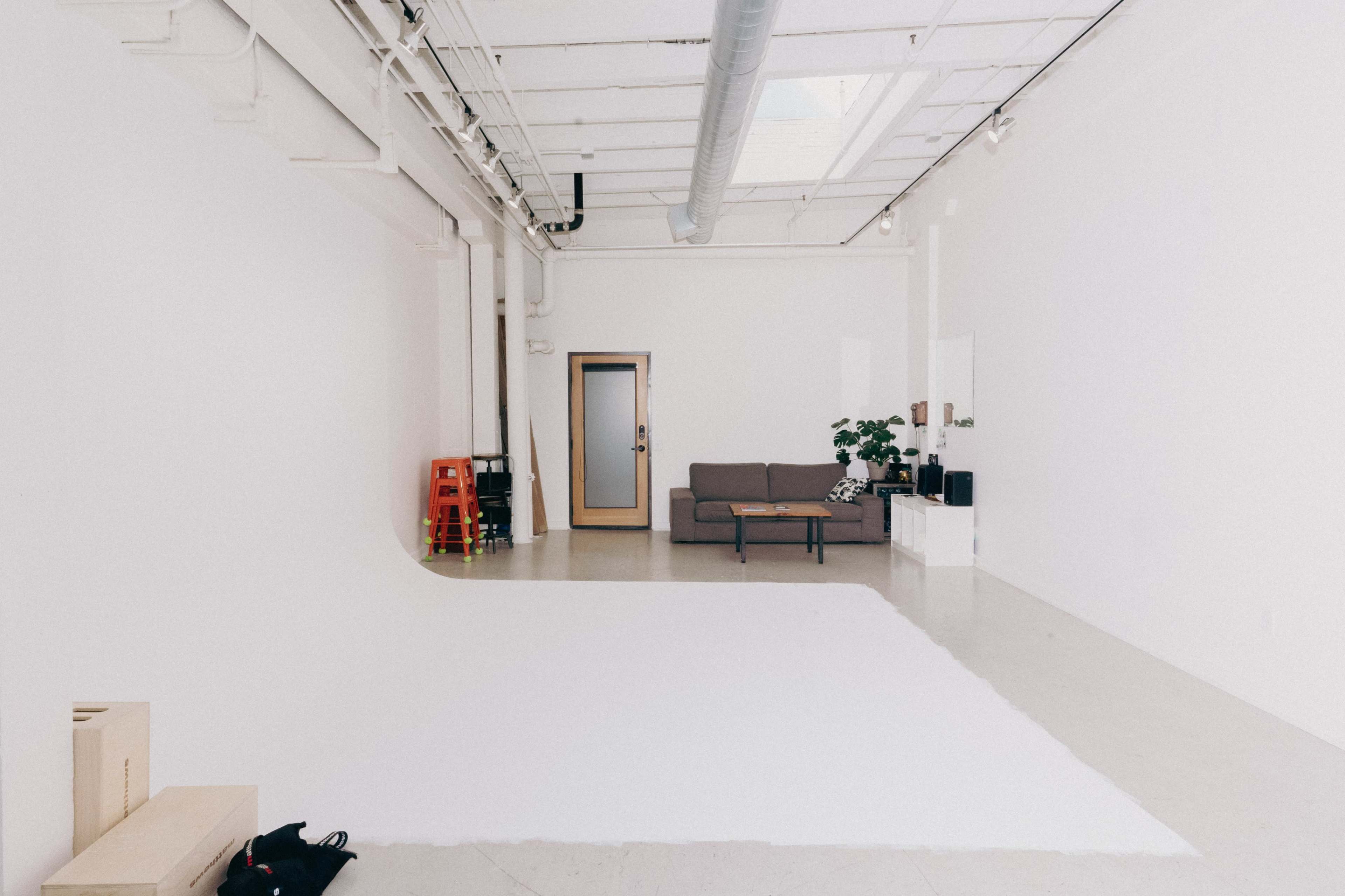 A minimalist, bright studio space featuring a gray sofa, a small plant, and a large white area on the floor.