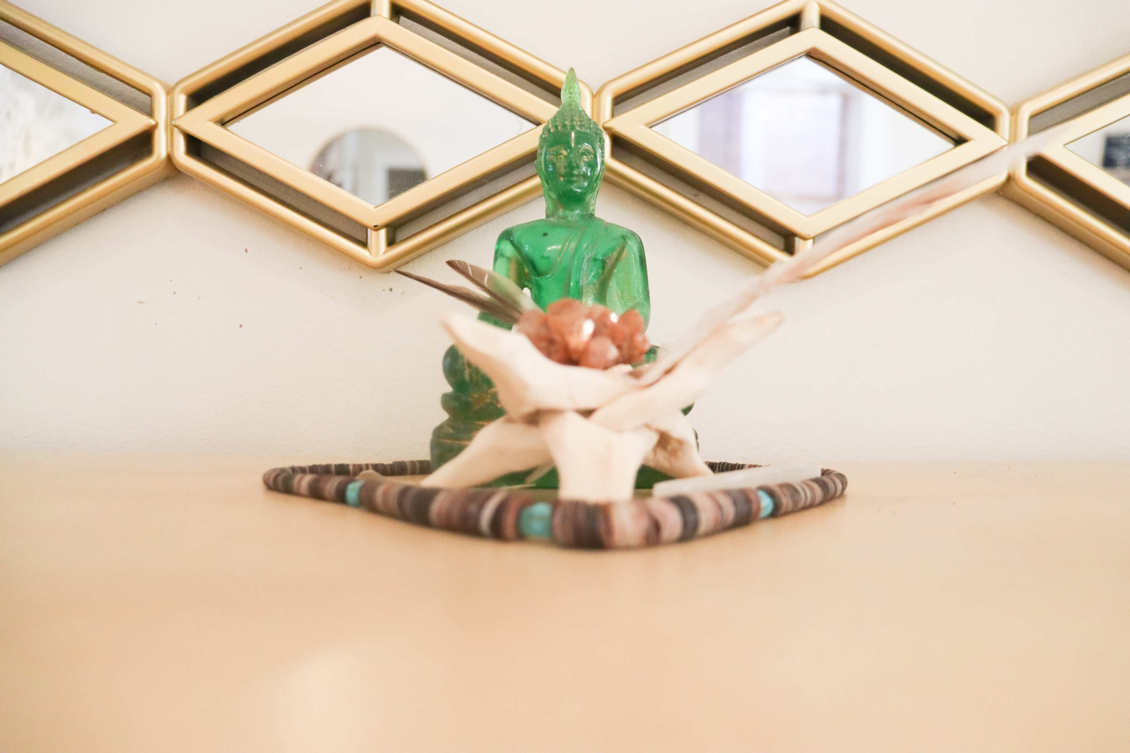 A green Buddha statue is placed atop a decorative arrangement of natural materials on a wooden surface, set against a backdrop of geometric mirrors.