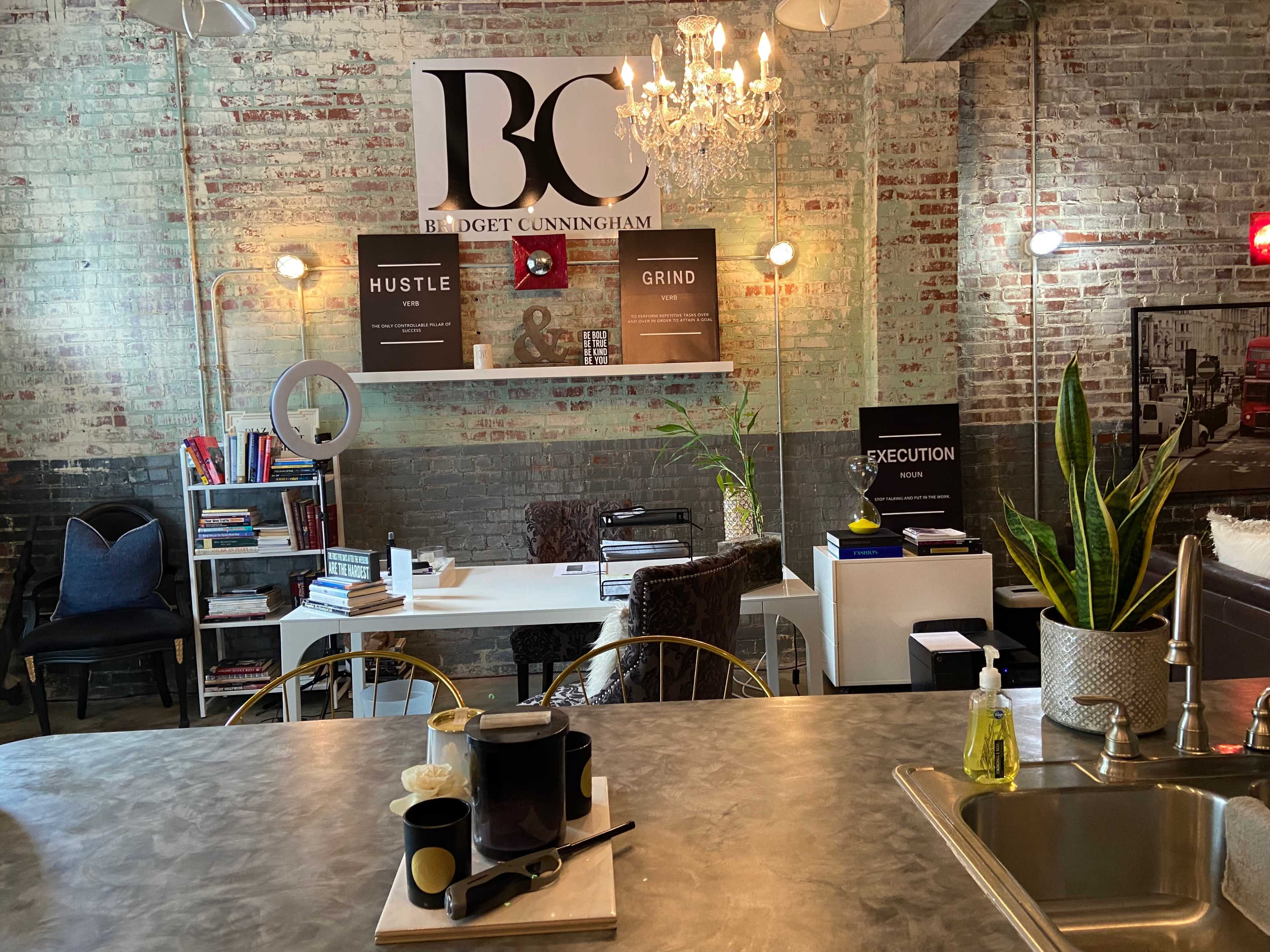 A modern office space with brick walls, a central work area featuring a desk and bookshelves, and a stylish chandelier overhead.