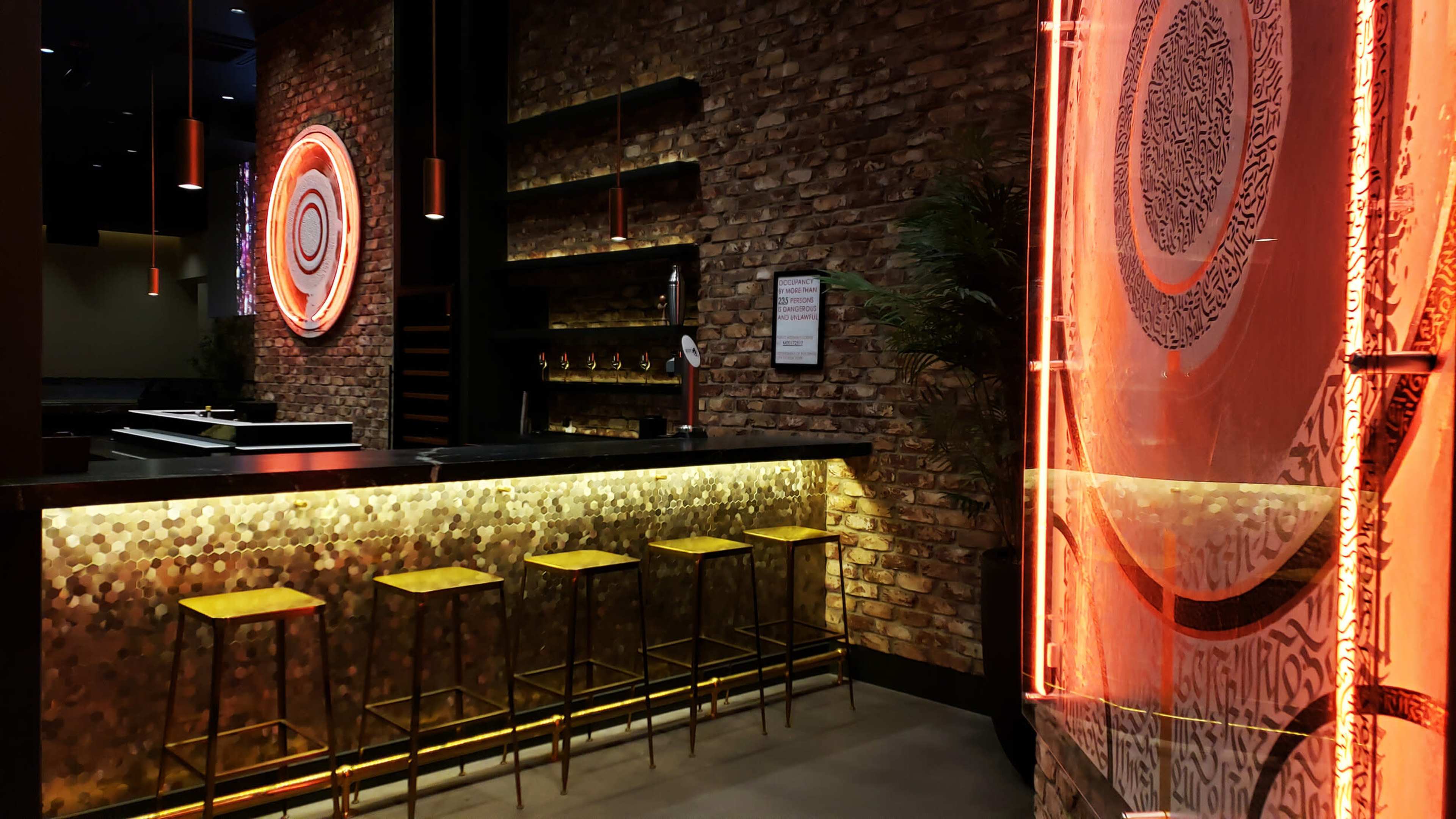 A modern bar area with a textured brick wall, glowing amber under-bar lighting, and golden stools arranged along a dark countertop.