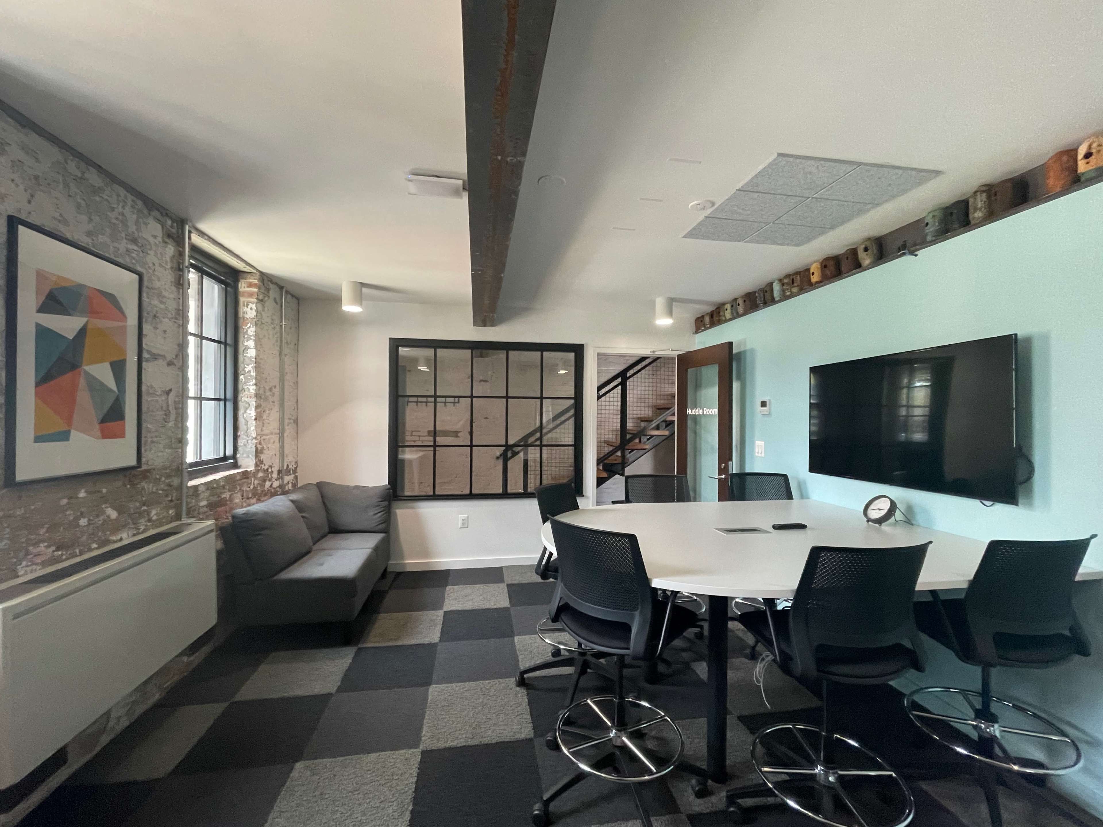 A modern meeting room features a gray couch, a table with high-backed chairs, large windows, and an adjacent staircase.