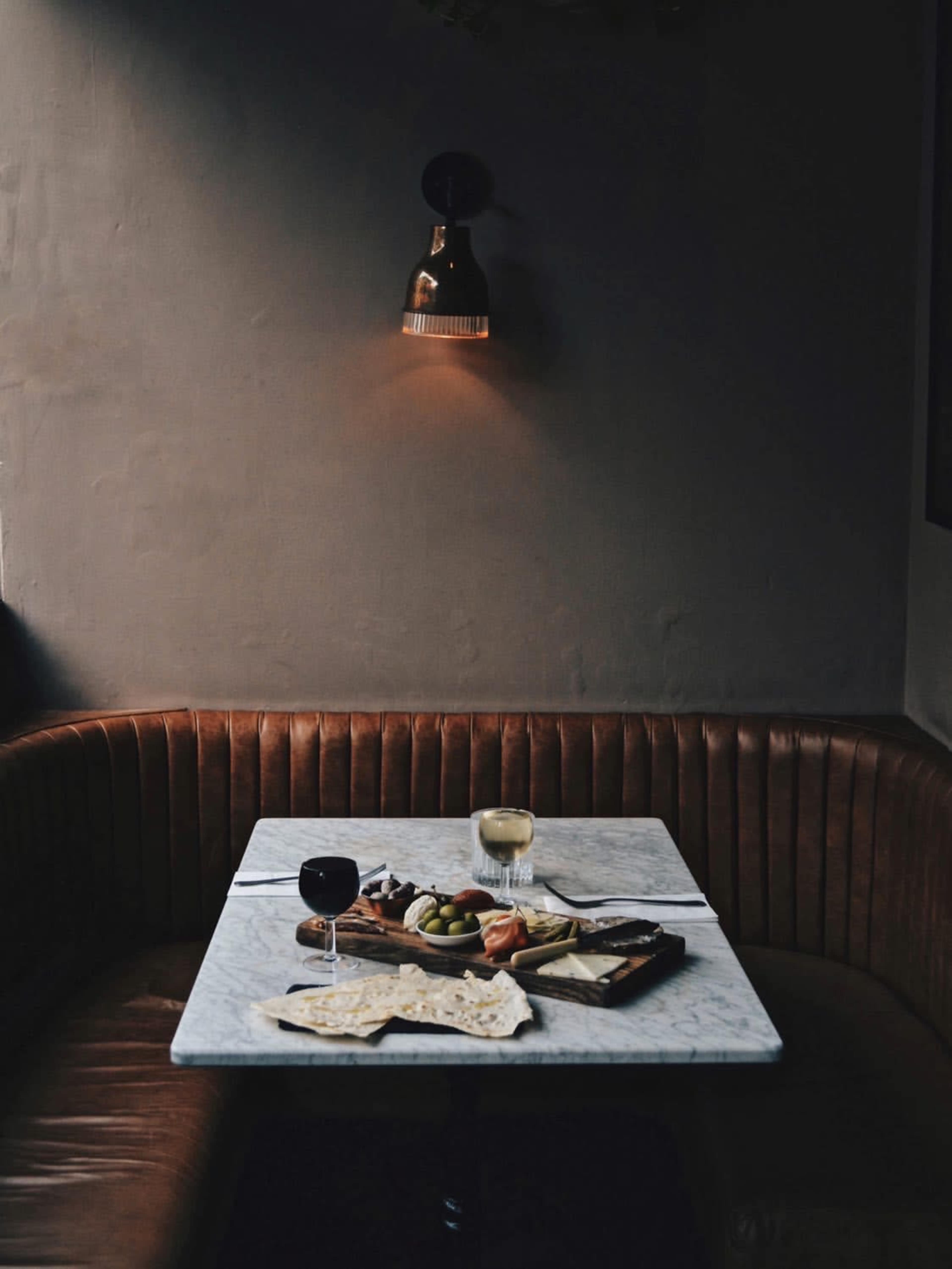 A table with a marble top is set with plates of food, glasses of wine, and a brown leather banquette in a dimly lit restaurant.