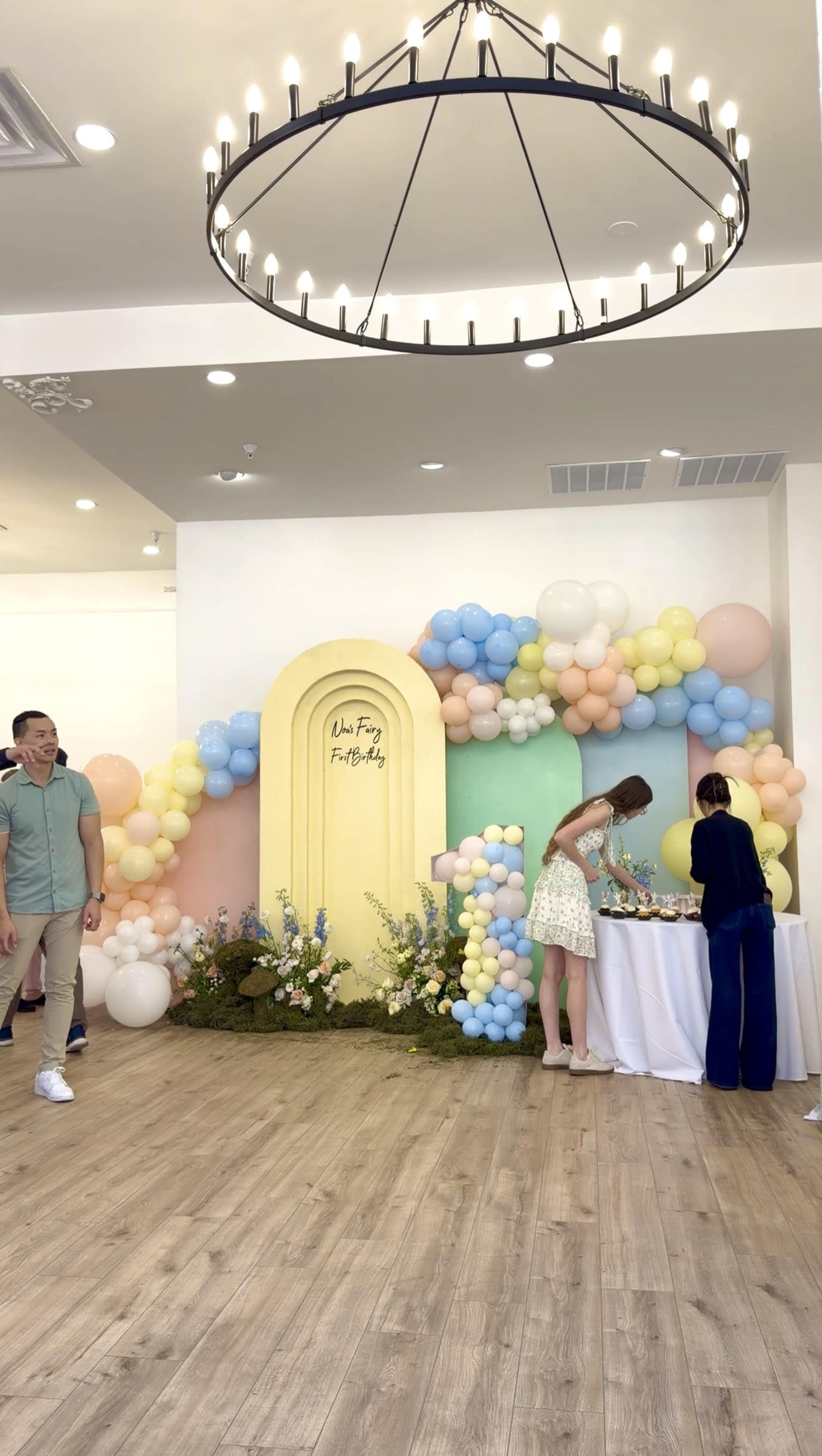 A festive indoor space is decorated with colorful balloons and flowers, featuring a large archway in the background and people setting up a table.