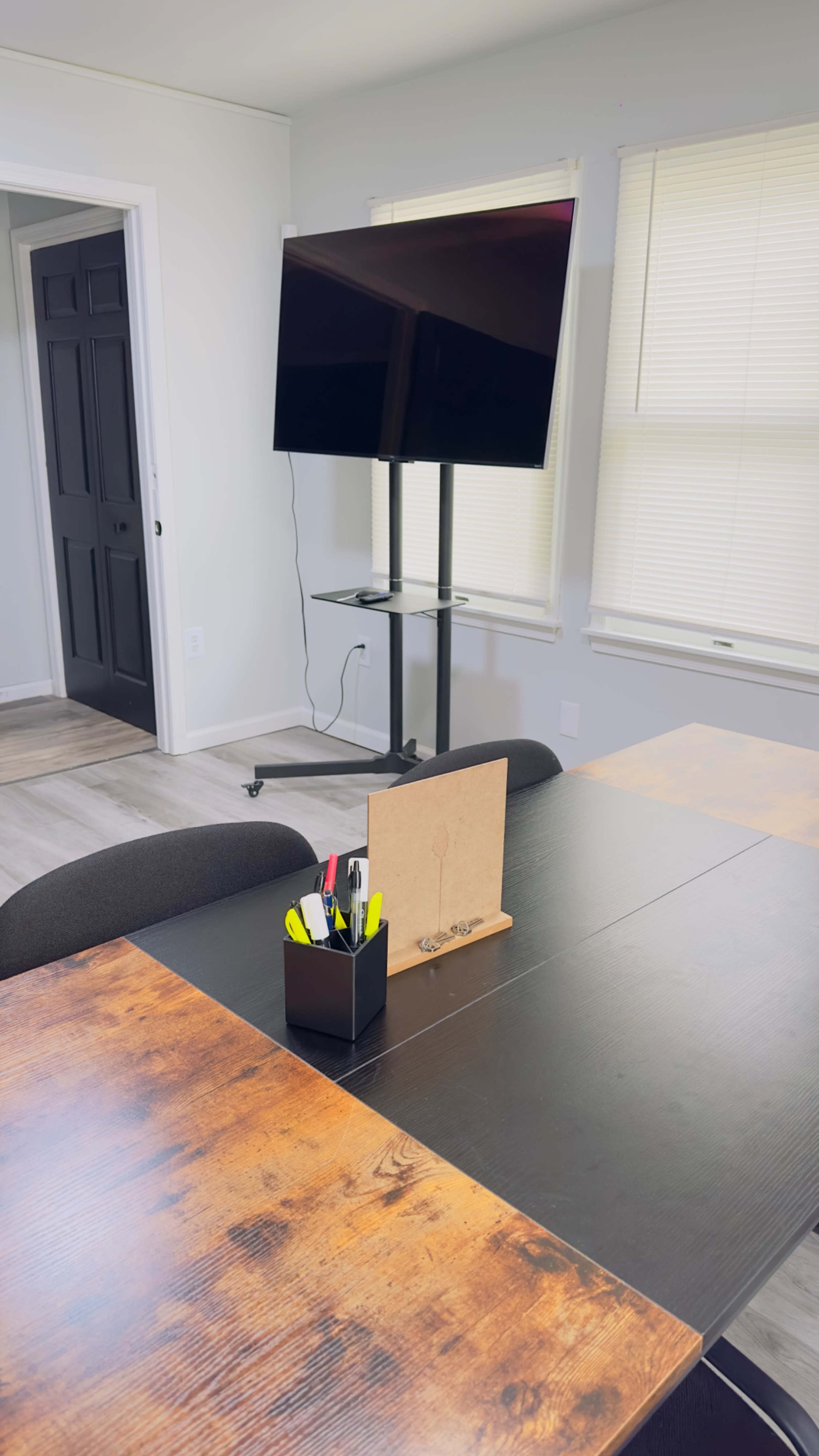 A conference room features a wooden table, a black pen holder, and a mounted TV on a stand against a light-colored wall.