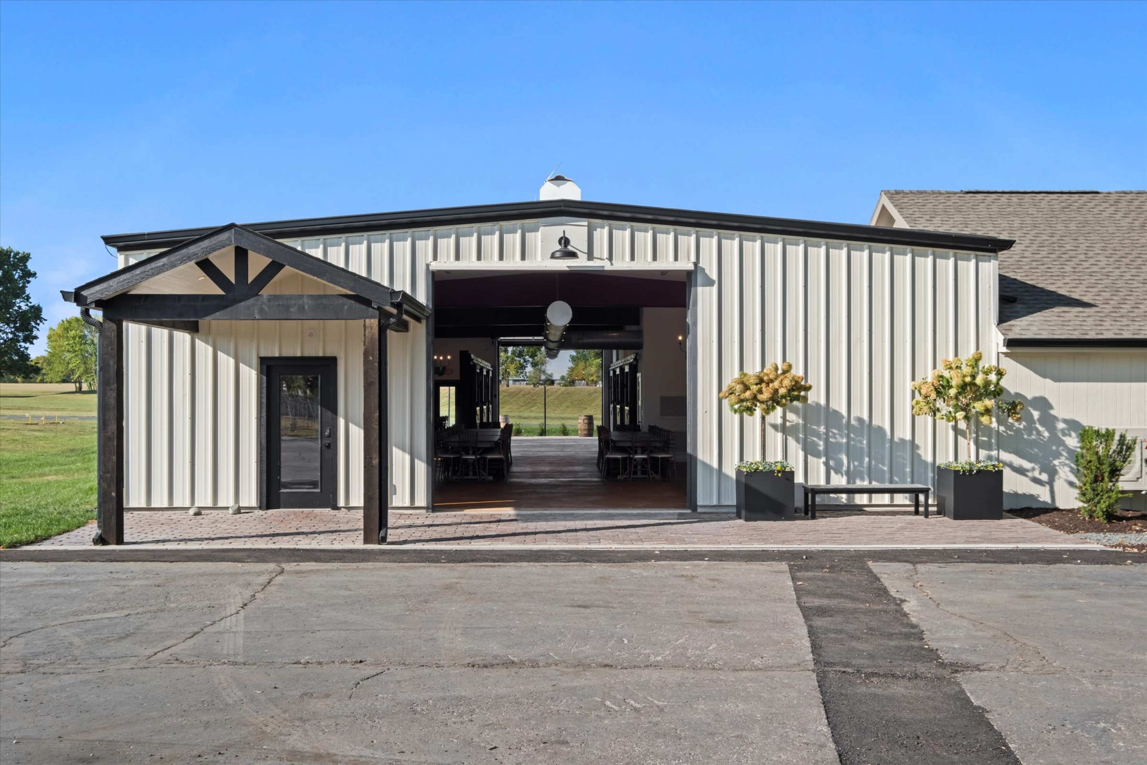 A modern building with a metal exterior features a covered entrance and large open doors leading to an interior space, surrounded by greenery.