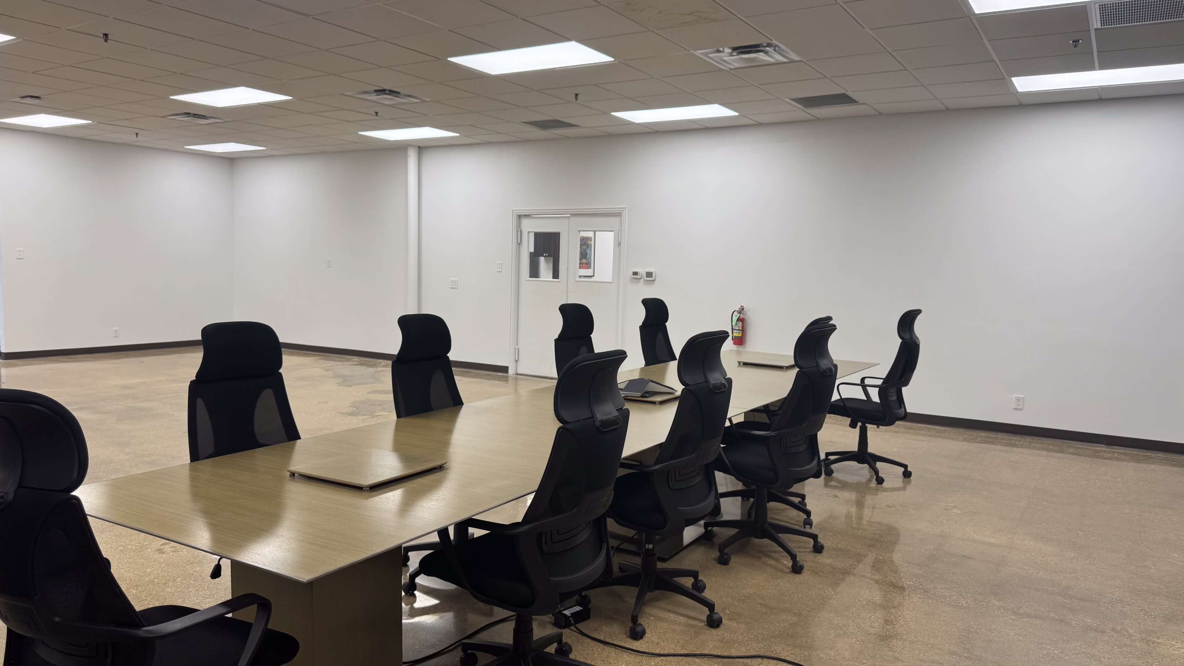 A long conference table with several black rolling chairs is positioned in a sparse, well-lit meeting room with white walls and a polished floor.