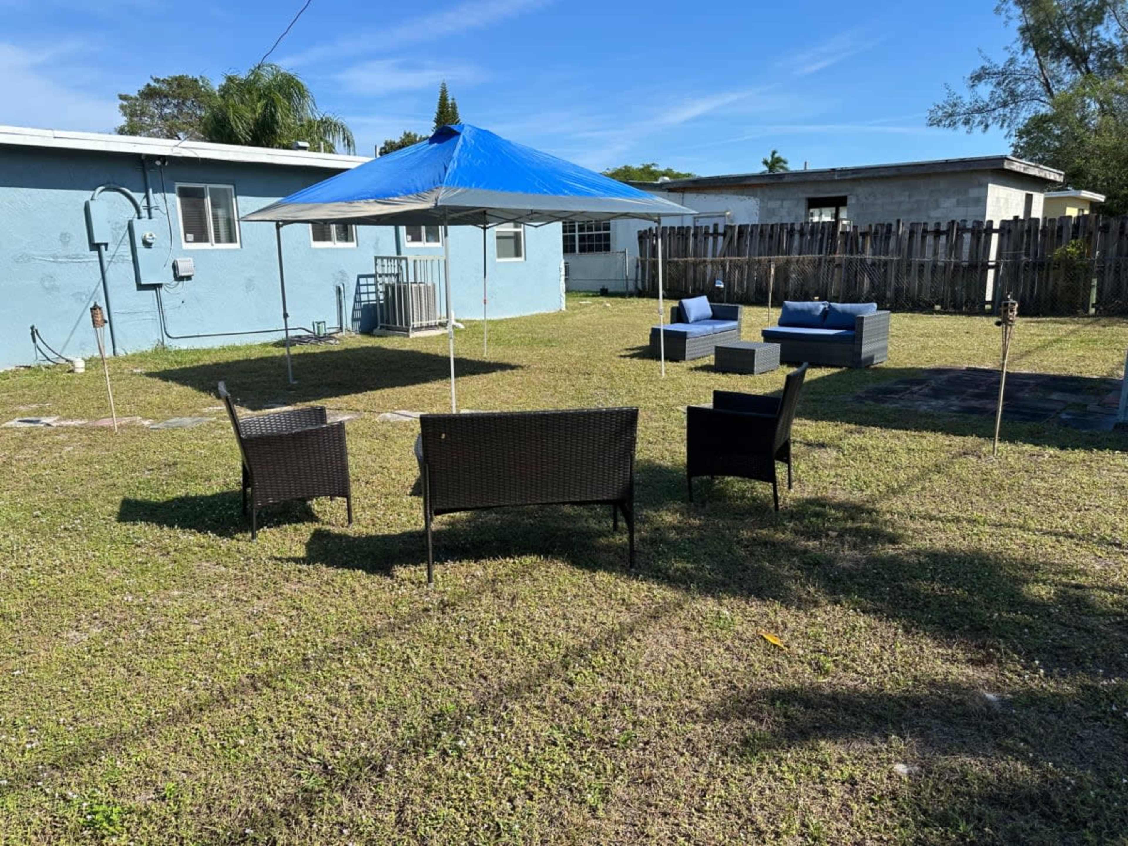 A backyard with a blue umbrella over a seating area, consisting of four dark wicker chairs and a large sofa, surrounded by grass and a fence.