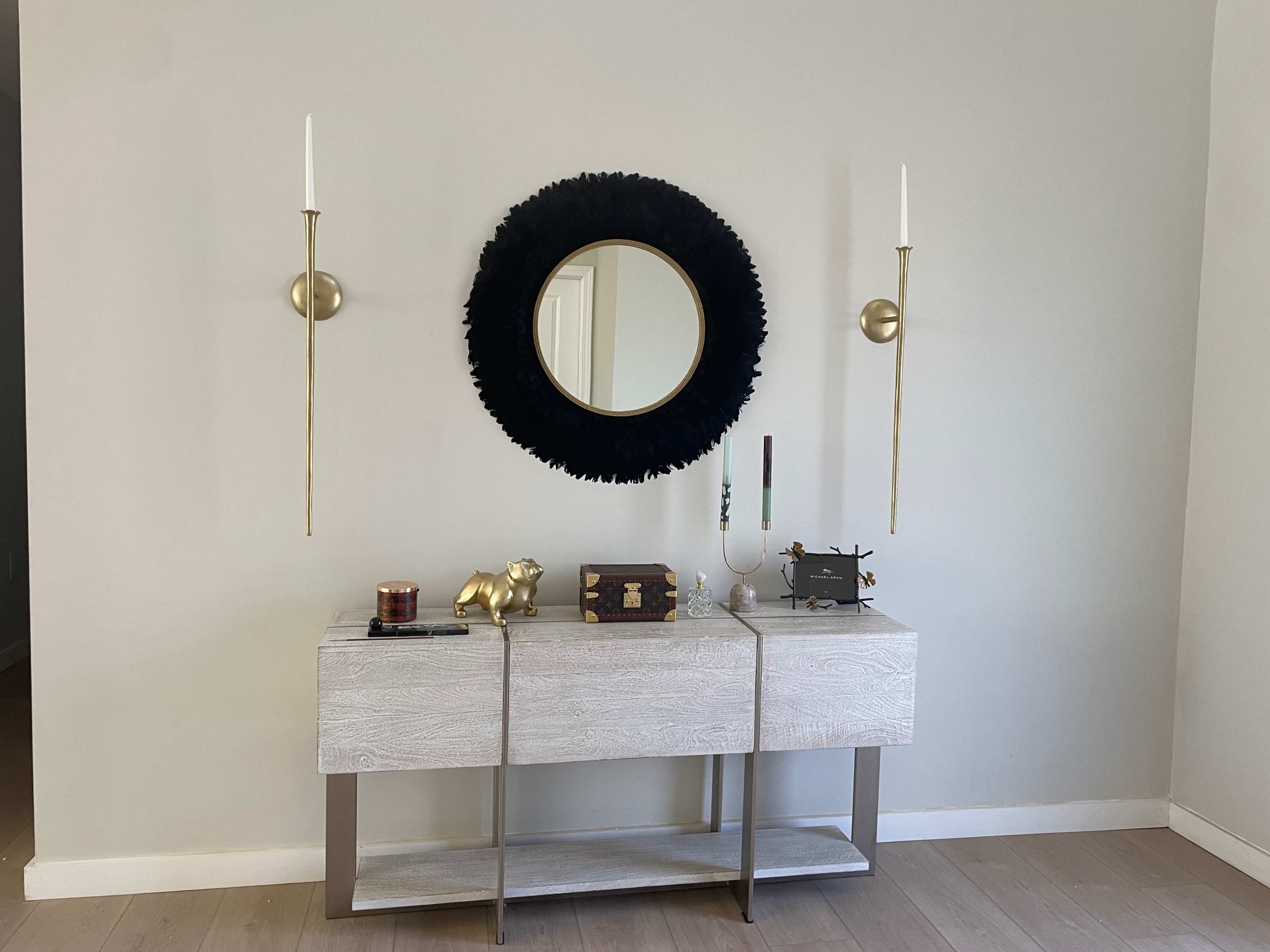 A dark circular mirror hangs above a light-colored console table, flanked by two gold wall sconces, with decorative items placed on the table.