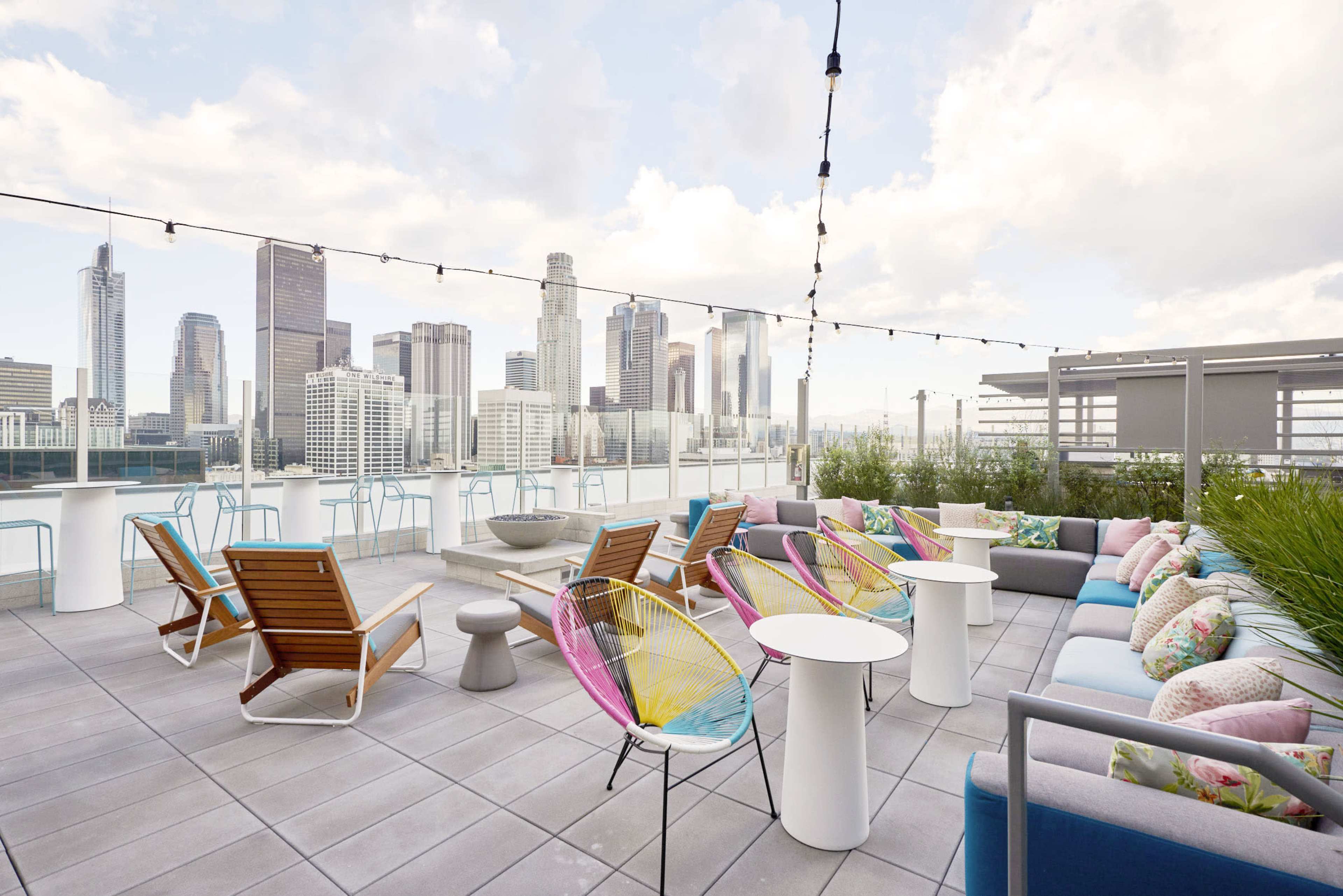 A rooftop lounge area features colorful seating, tables, and panoramic views of a city skyline under a cloudy sky.
