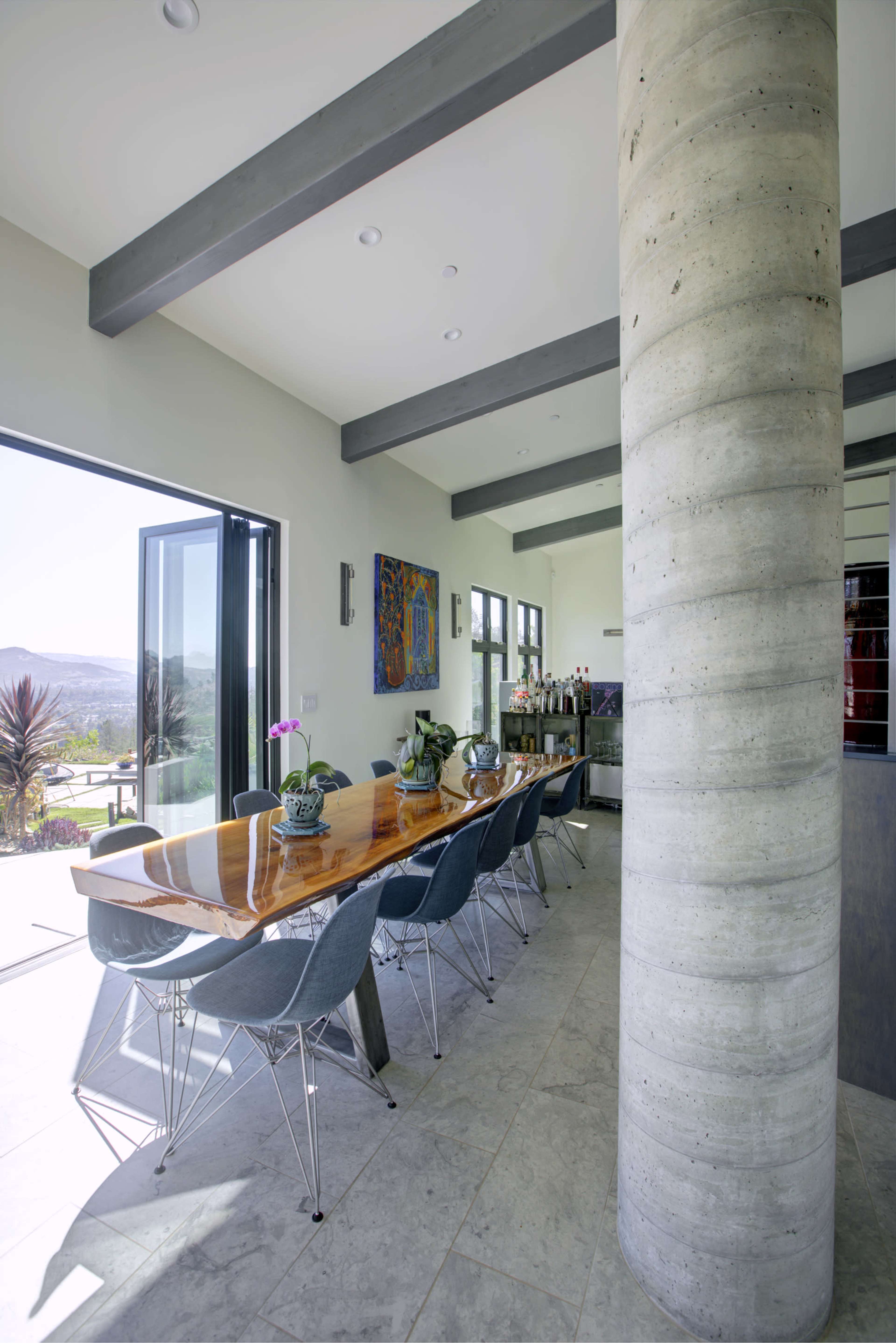A long wooden dining table with gray chairs is positioned near a large opening that leads to an outdoor view, with a concrete pillar supporting the ceiling.