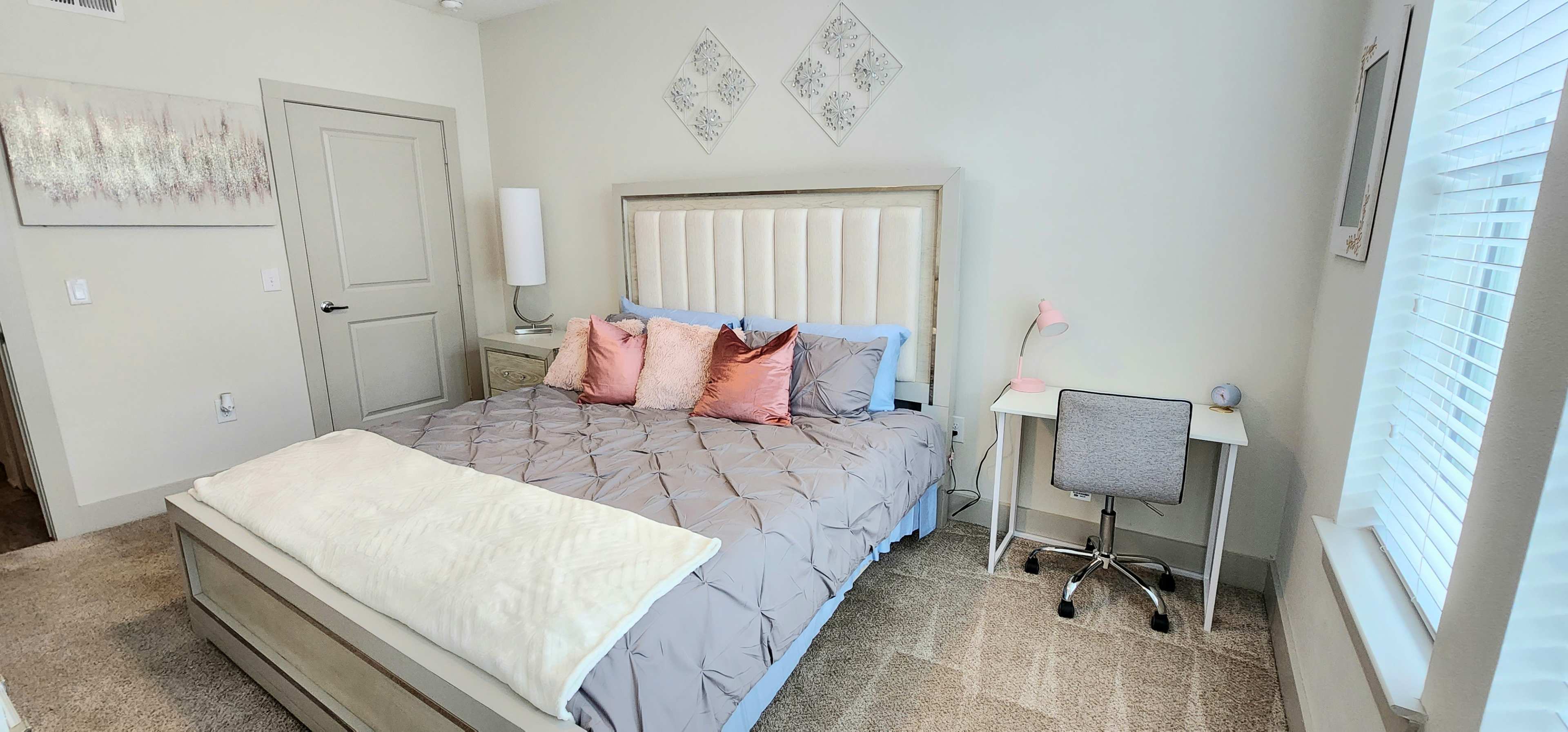 A neatly arranged bedroom featuring a large bed with a light gray coverlet, decorative pillows, a nightstand, a desk with a chair, and a window with blinds.