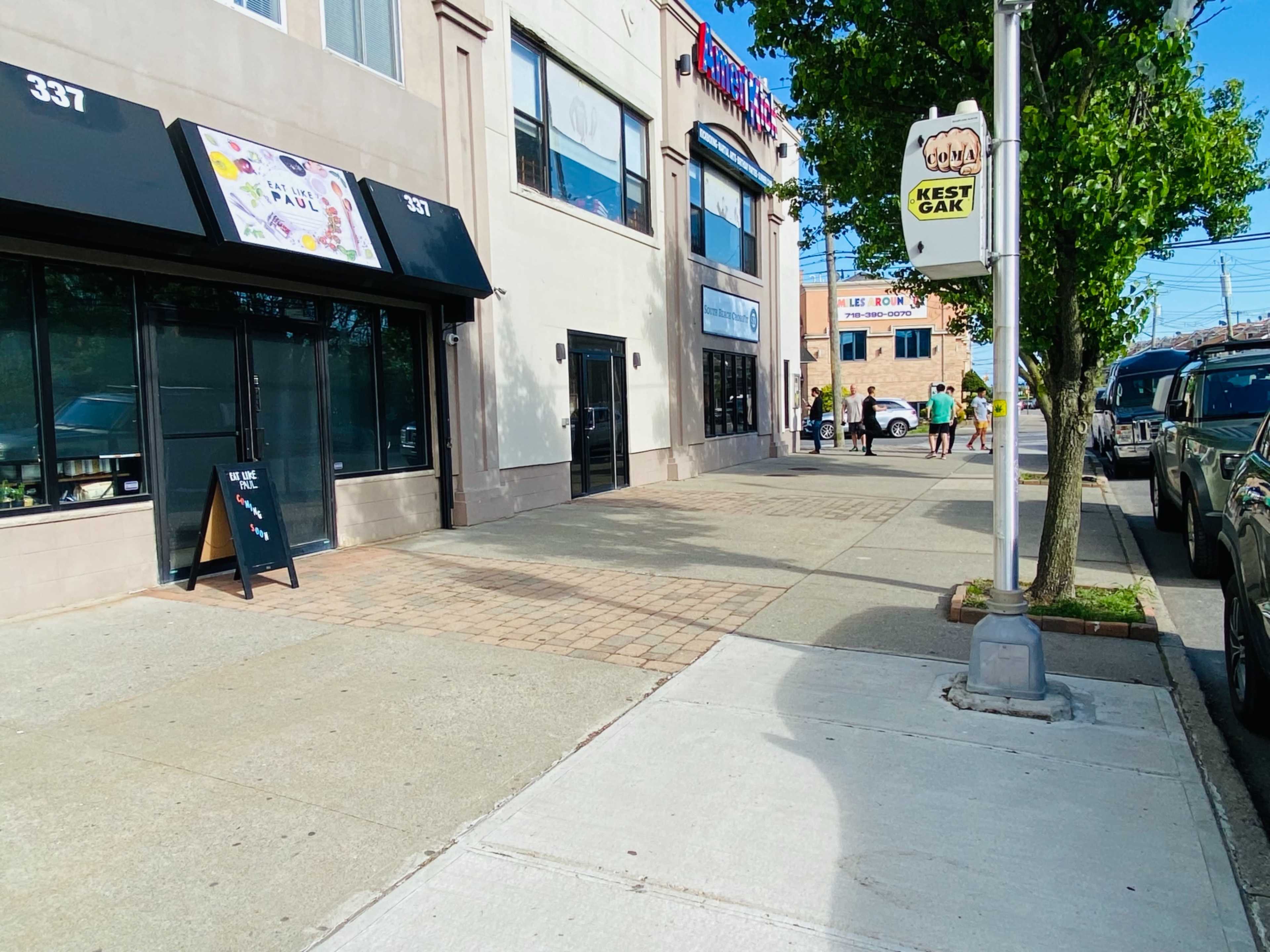 The image shows a street with a sidewalk lined by storefronts, parked cars, and a sign for a business visible on the right.