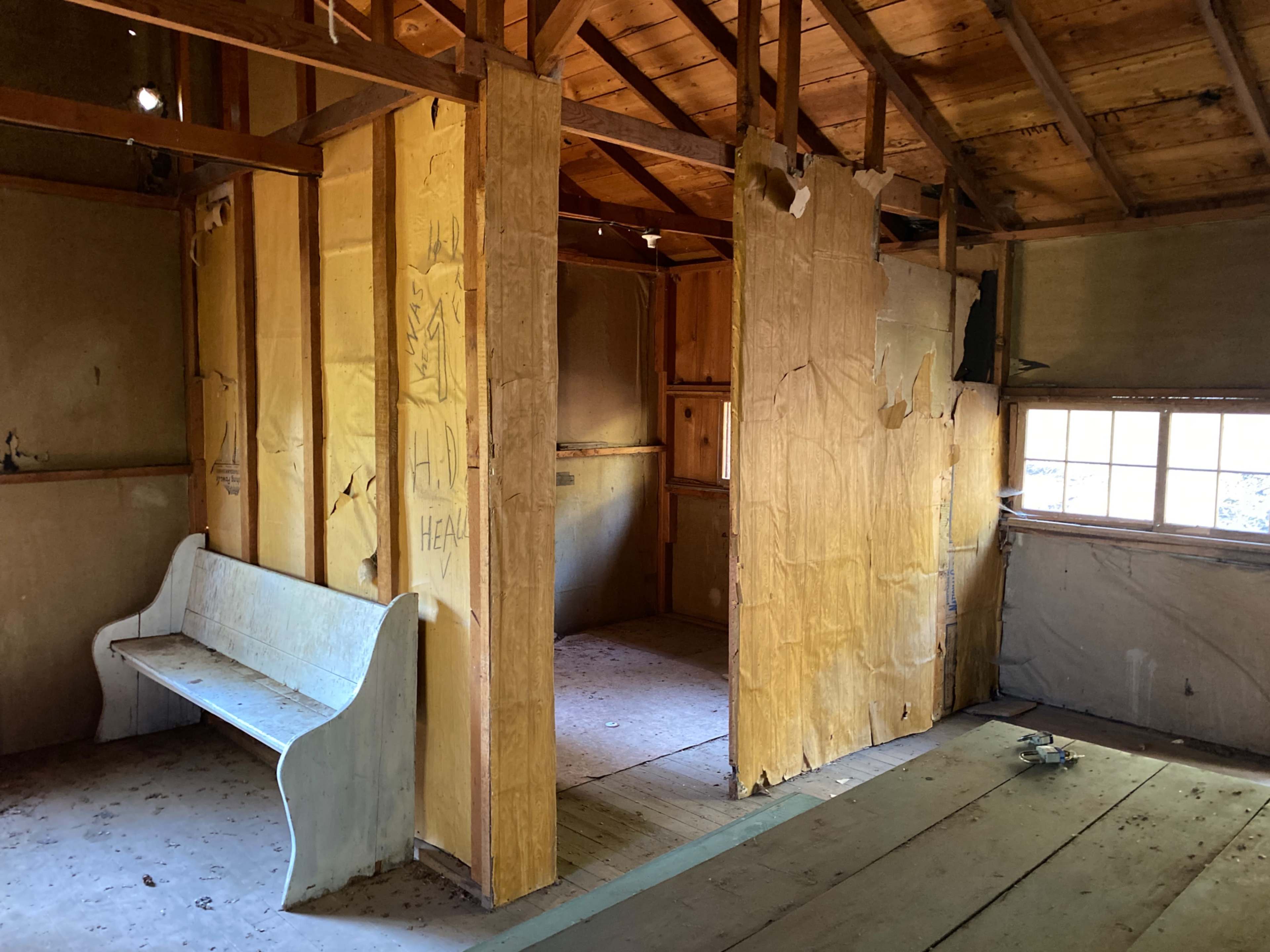 The interior of an abandoned building features exposed wooden beams, partially torn wall coverings, and a wooden bench against one wall.