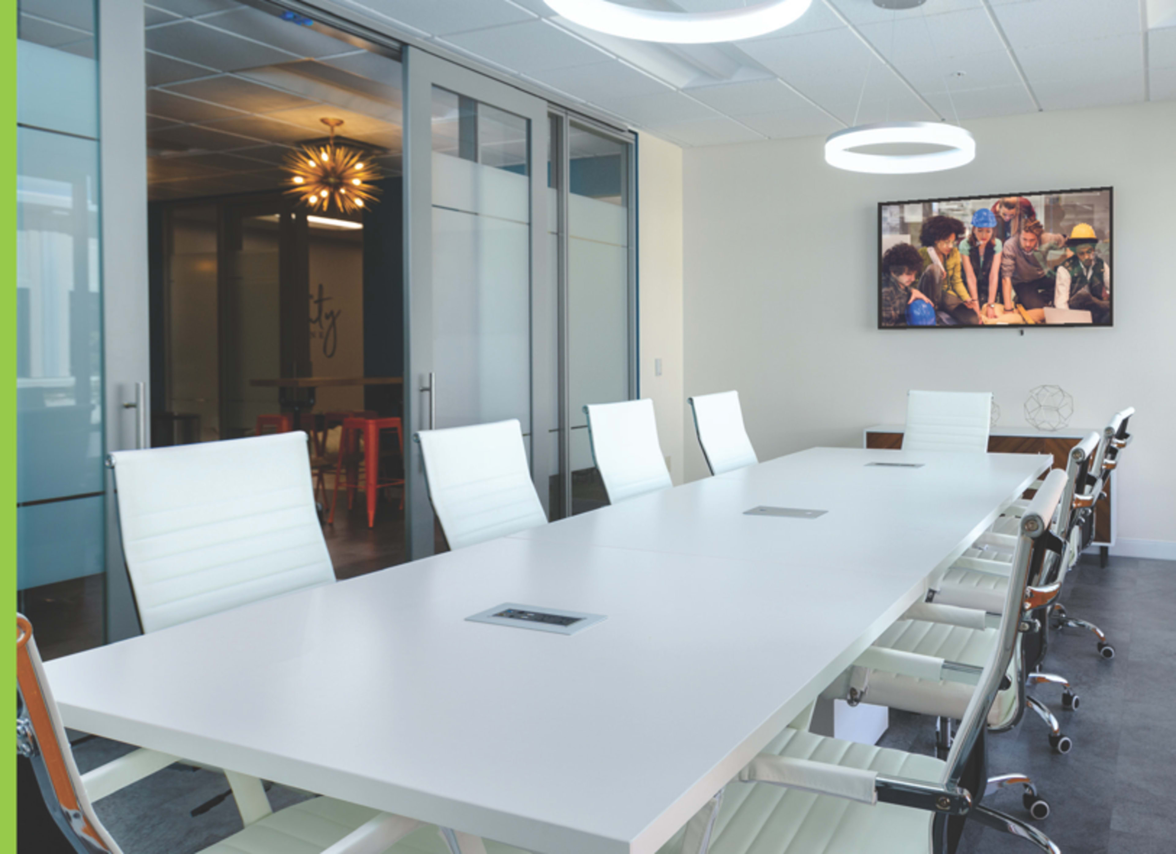 A modern conference room features a long white table surrounded by ergonomic chairs, with sliding glass doors and a television displaying a group of people collaborating.