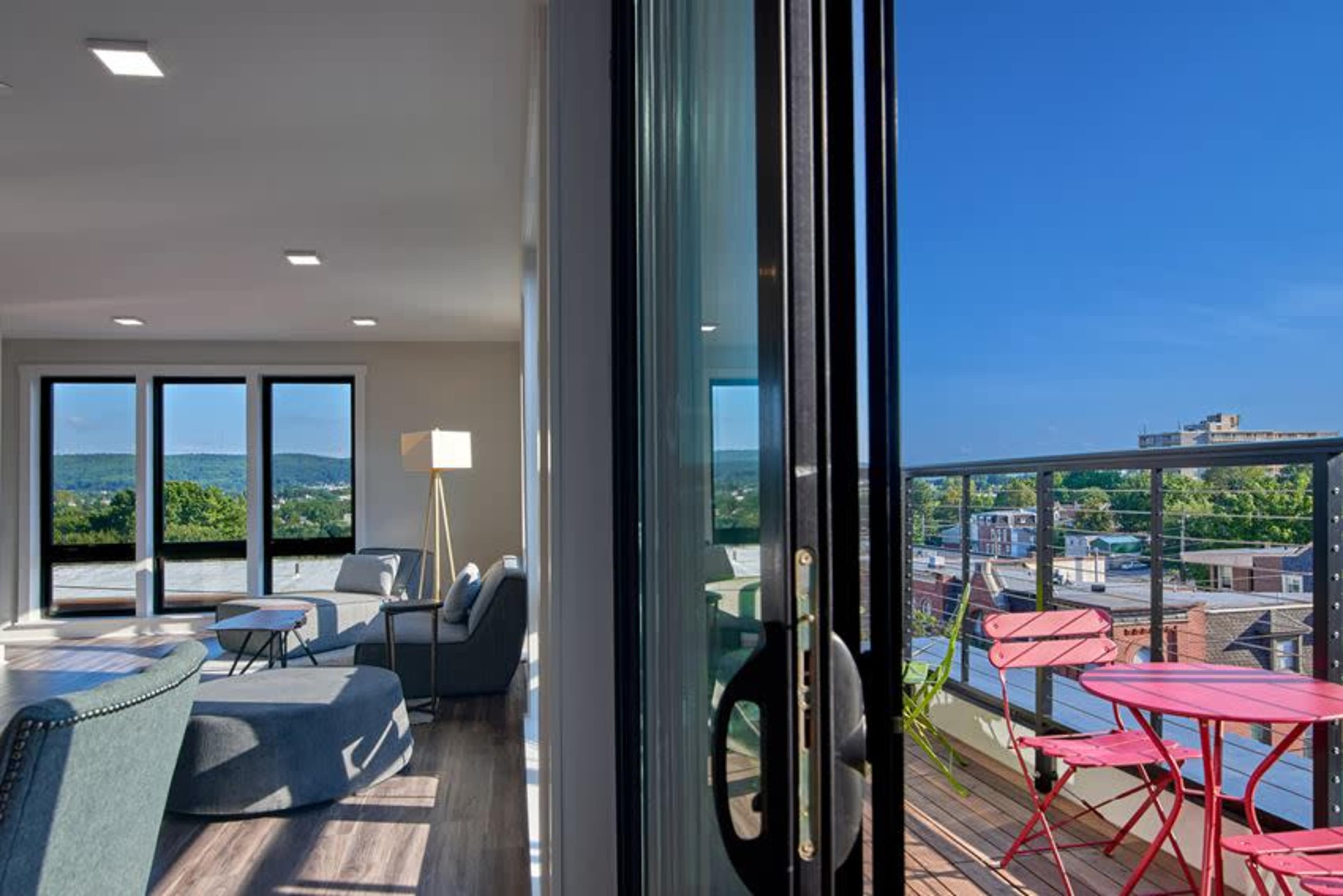 A modern living room with large windows opens onto a balcony featuring a small table and chairs, overlooking a cityscape against a clear blue sky.