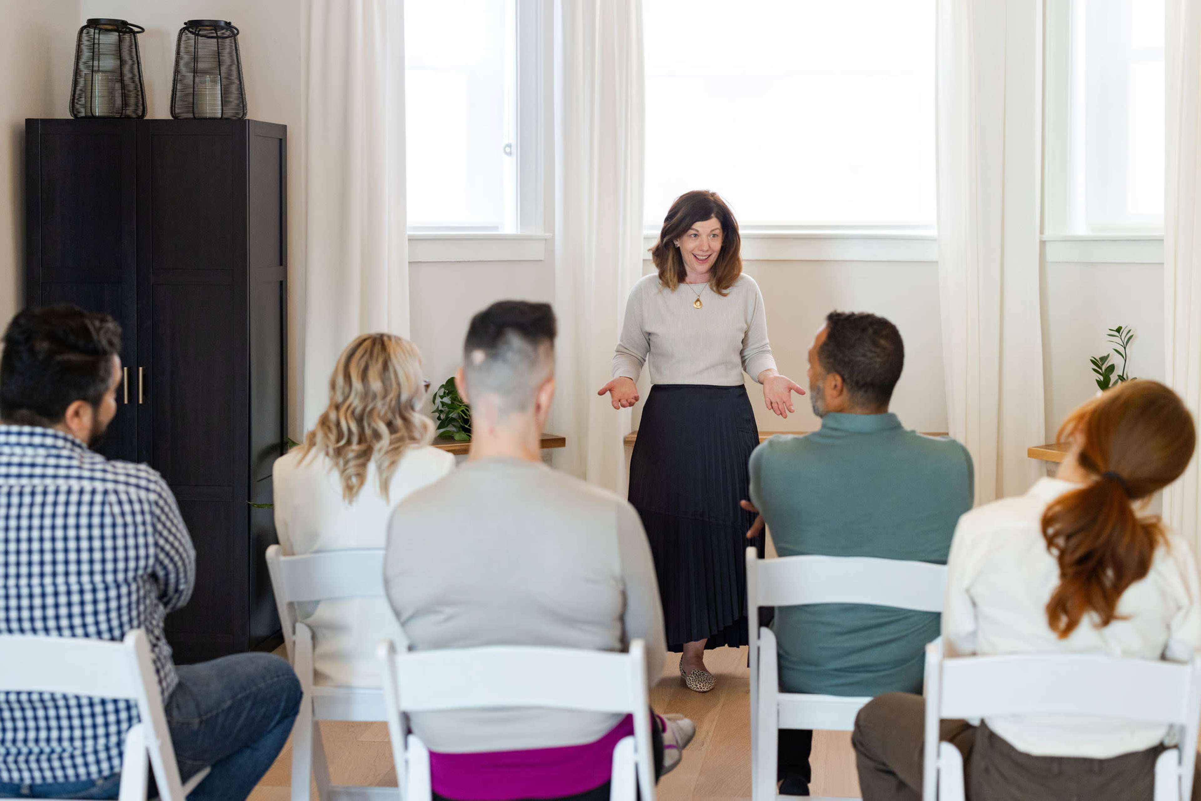 A speaker addresses an audience seated in white chairs in a well-lit room with plants and a dark cabinet in the background.