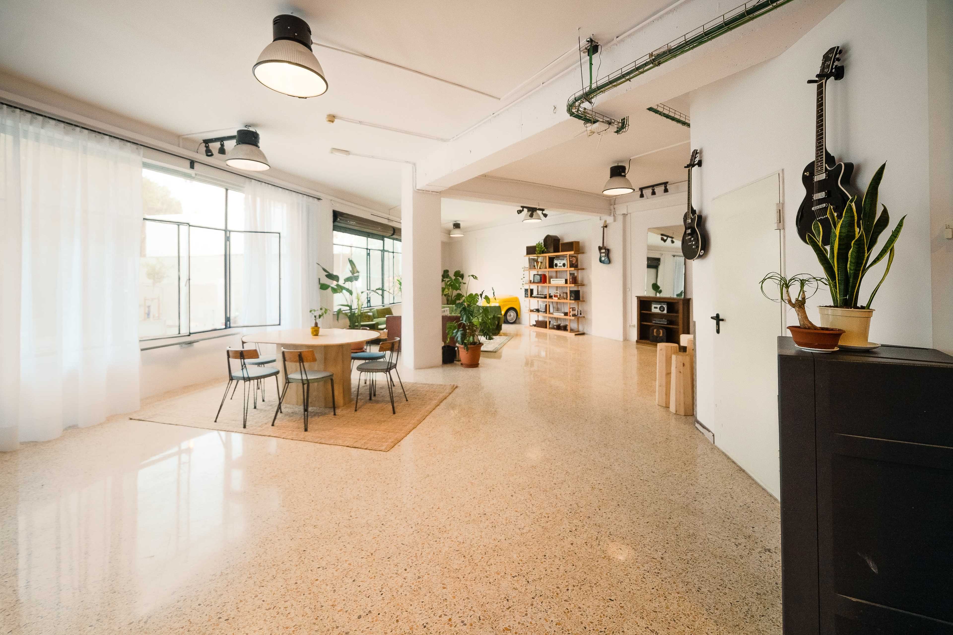 A spacious room with a dining table, plants, and a bookshelf, featuring large windows that allow natural light to enter.