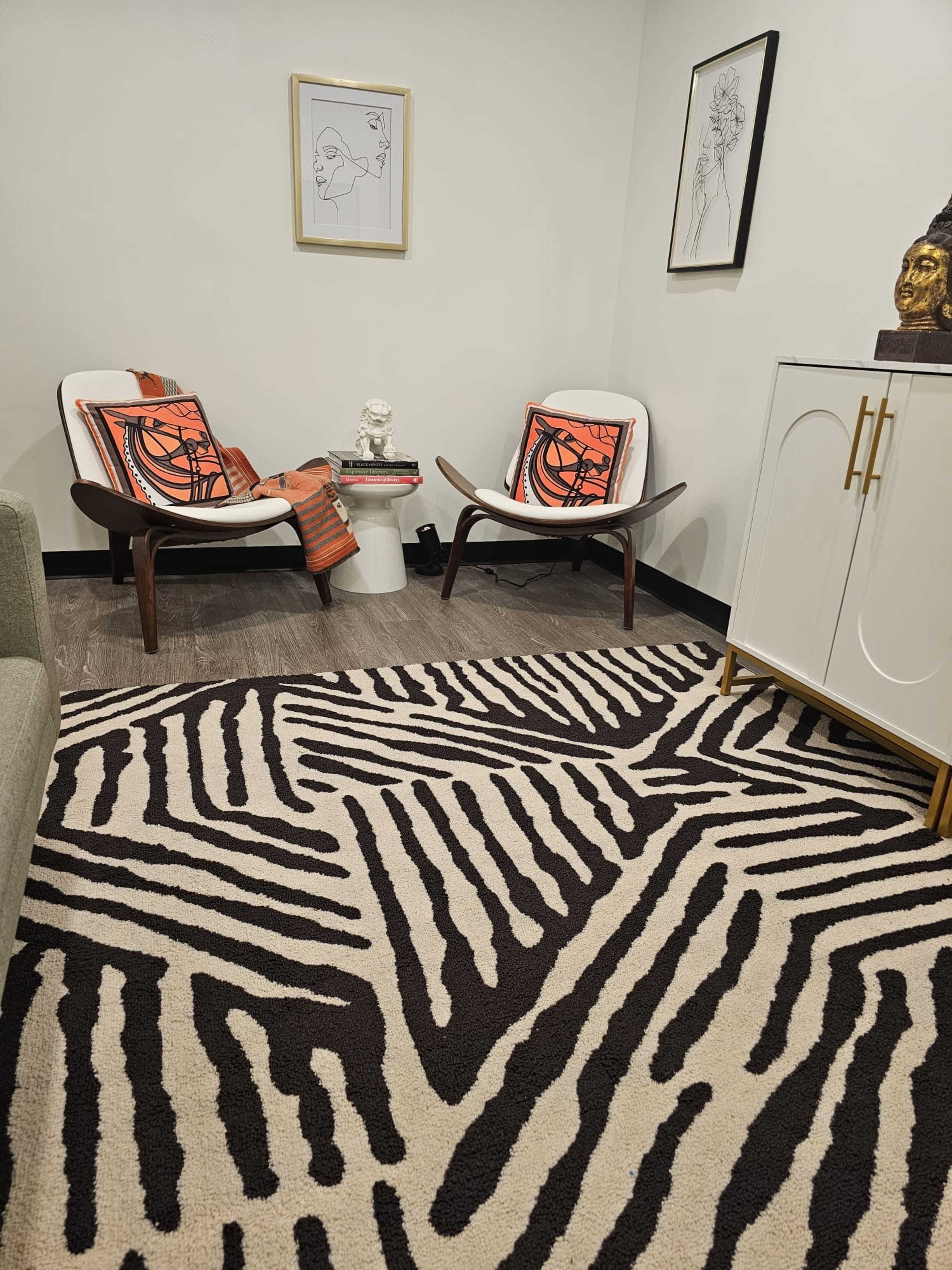 A cozy seating area features two modern chairs with orange cushions, a small side table, and a zebra-striped rug, complemented by wall art and a decorative bust.