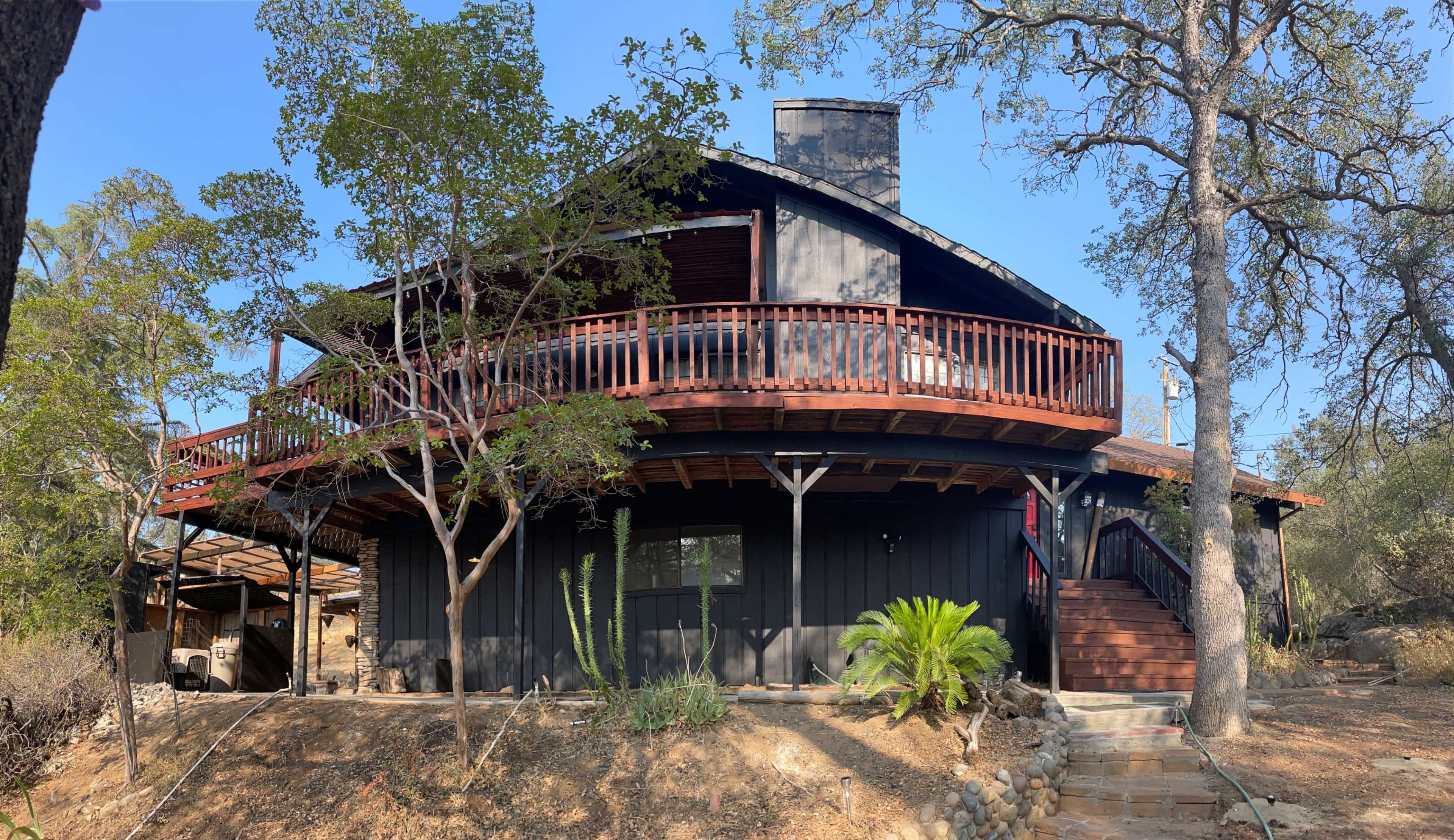 A modern, circular wooden house with a balcony, surrounded by trees and a landscaped area.