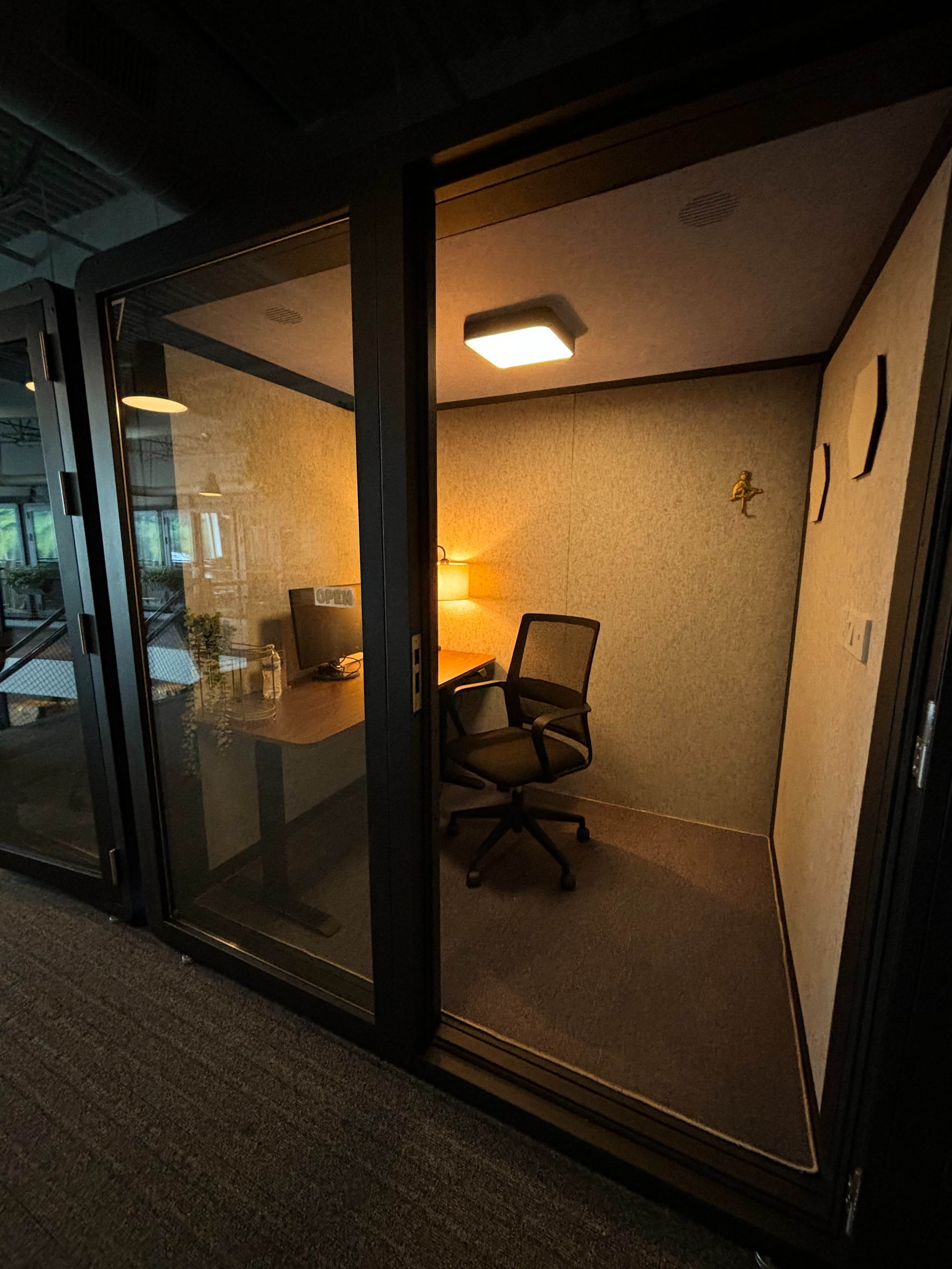 The image shows a small, enclosed office space with a desk, chair, and a lamp, all surrounded by glass walls.