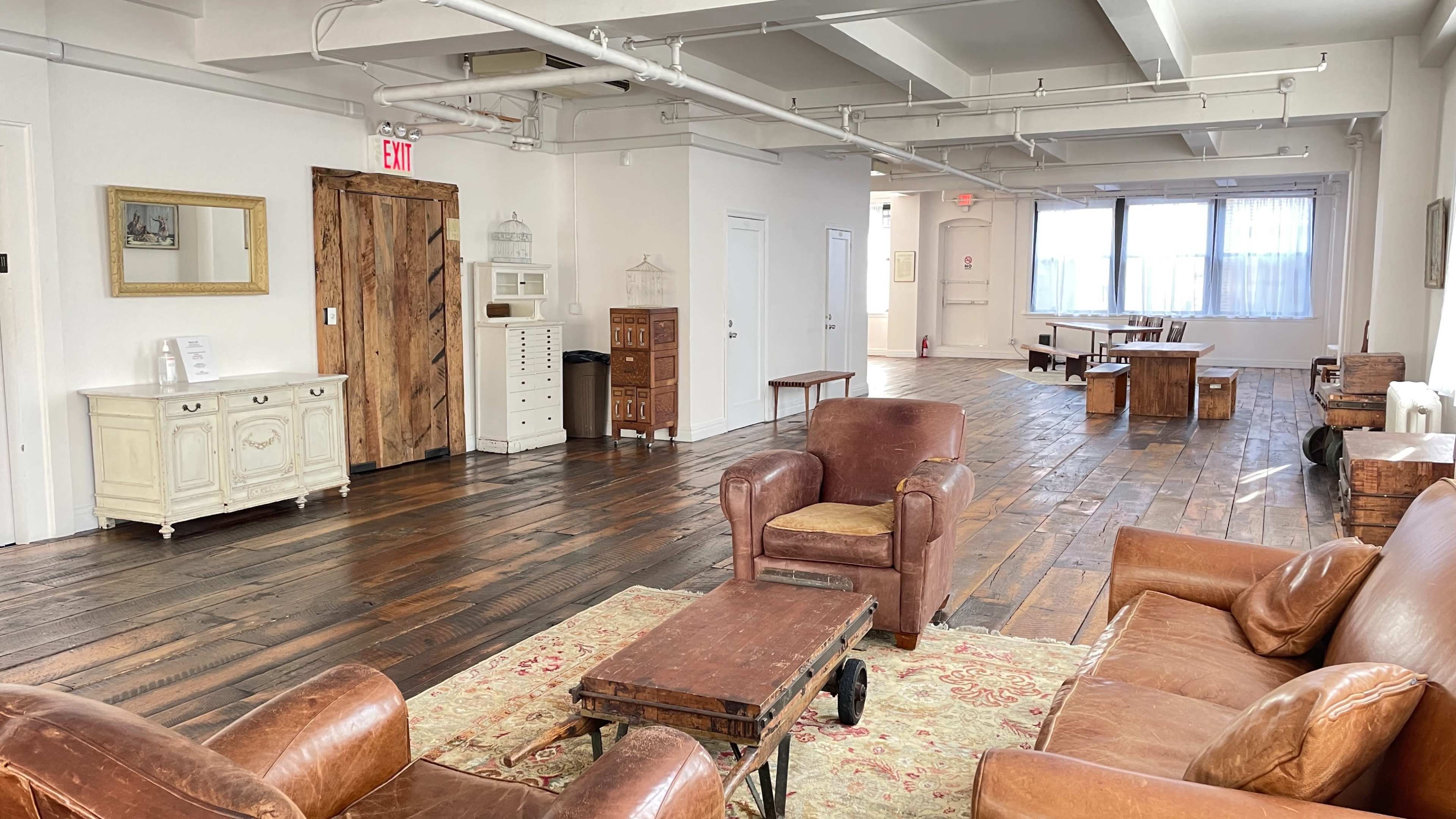 A spacious, well-lit room with wooden floors, featuring a seating area with brown leather chairs, a coffee table, and various wooden furniture pieces along the walls.