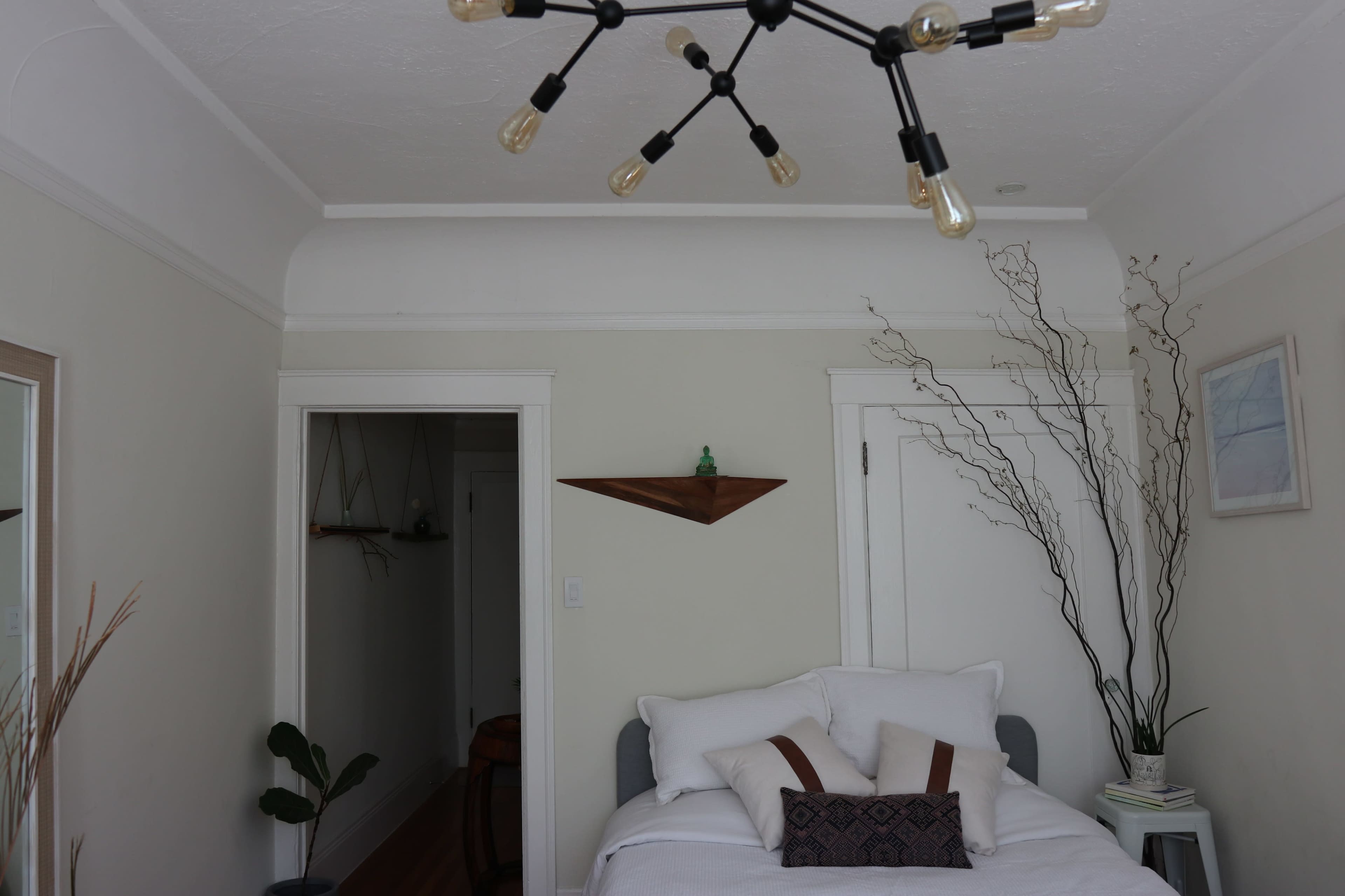 A bedroom with a bed, a ceiling light fixture, and decorative branches in a vase against a neutral-colored wall.