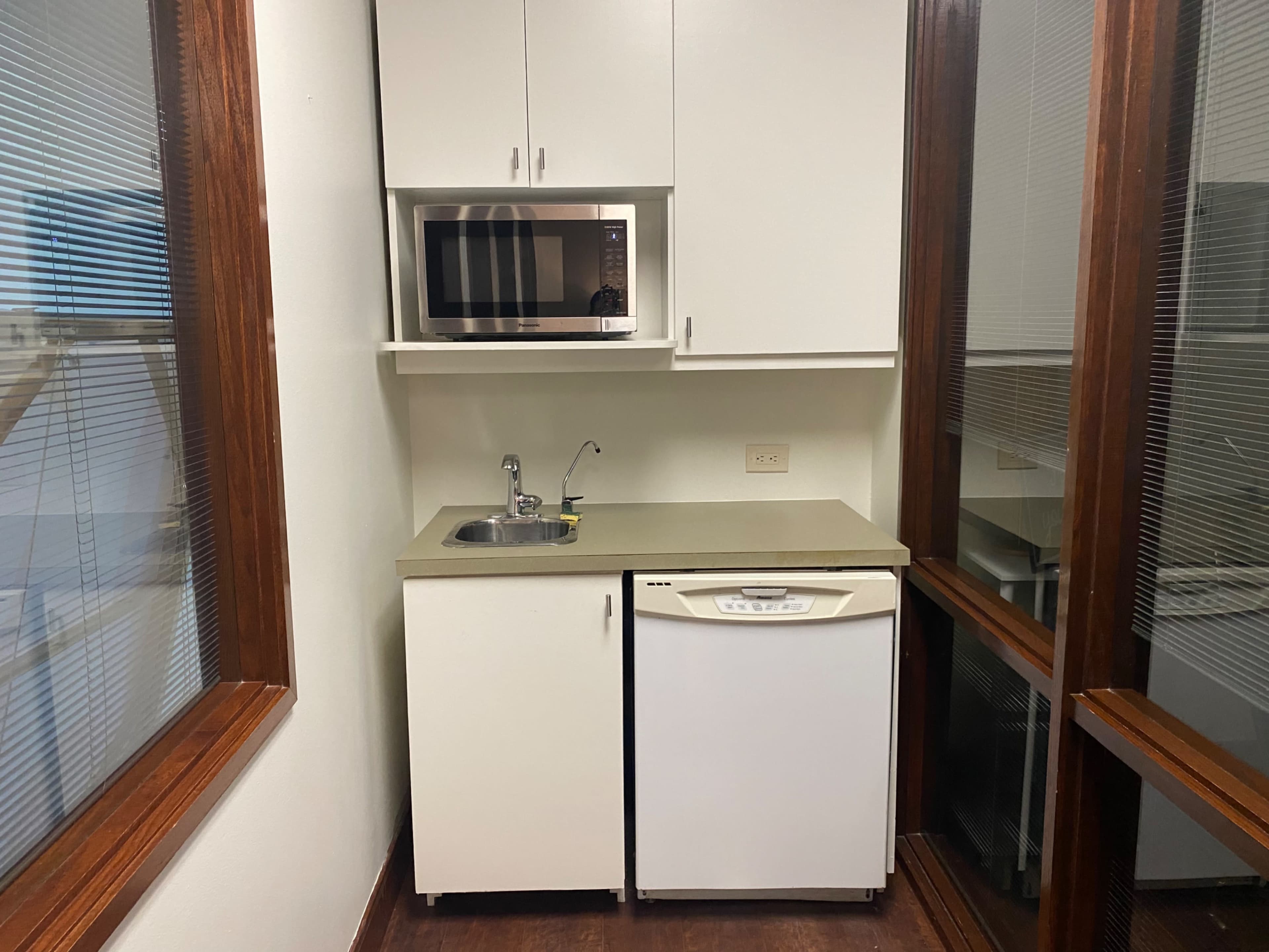 The image shows a small kitchen area with a microwave, sink, and refrigerator against a wall with windows.