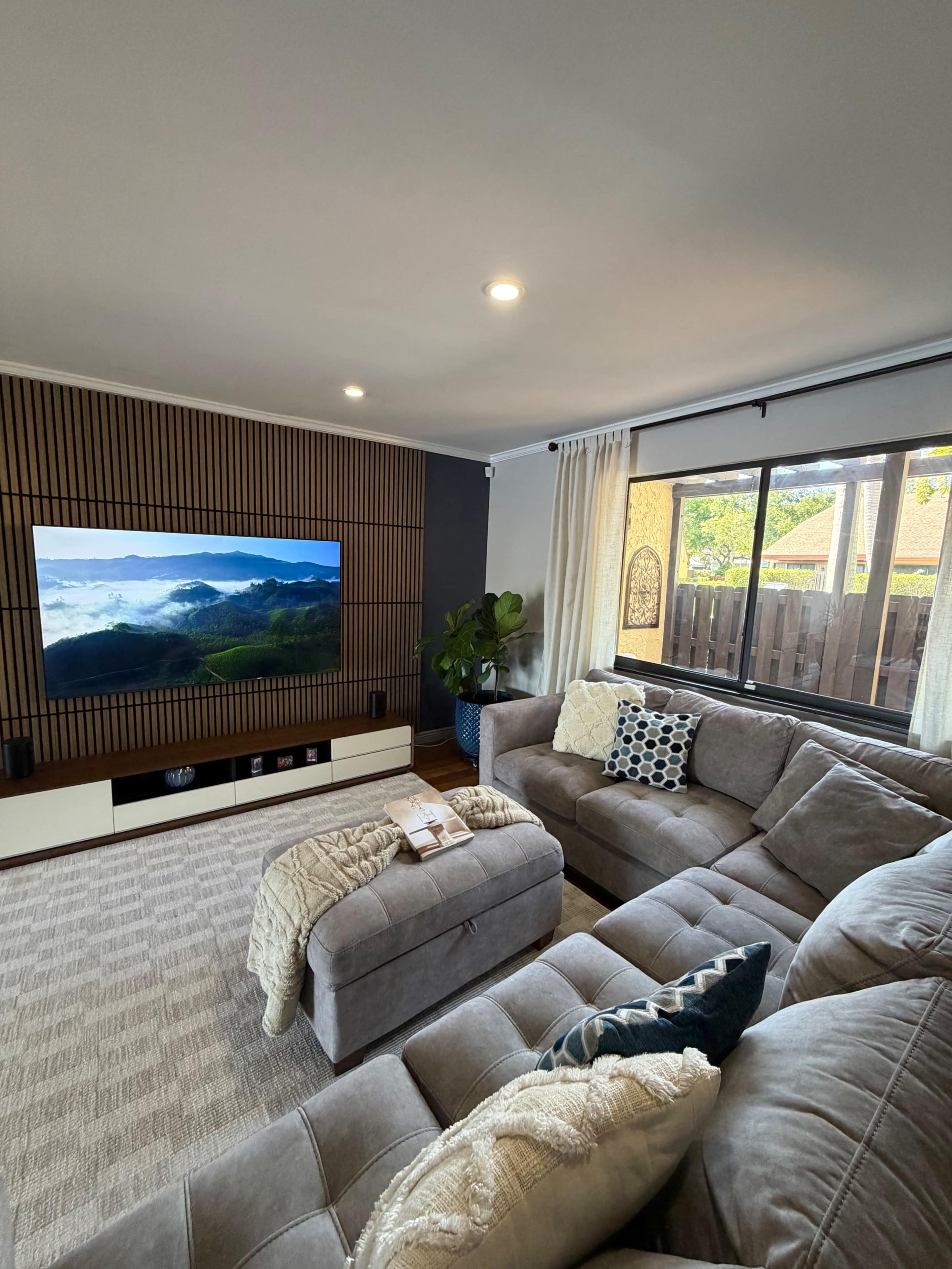 The image shows a modern living room featuring a large sectional sofa, a wall-mounted television, and a window with natural light.
