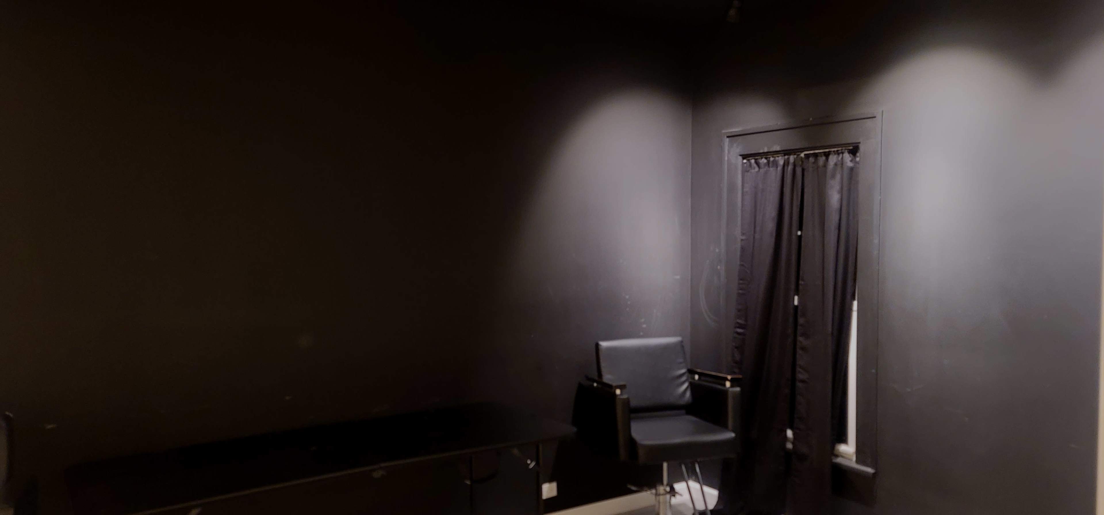 A dimly lit room with black walls, a black chair, and a window covered by a black curtain.